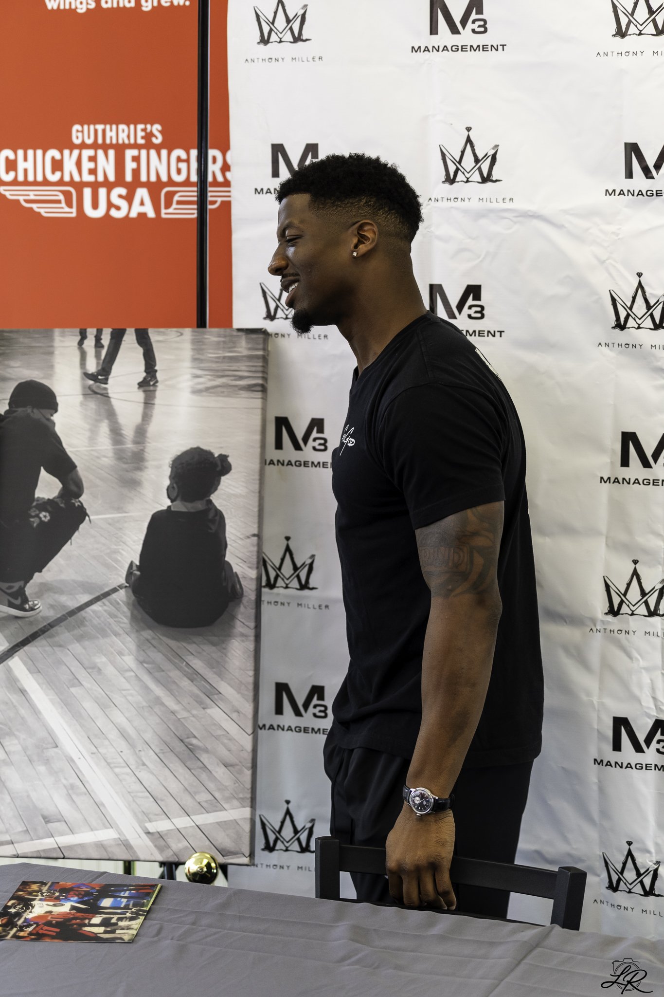 A man standing sideways at a table with photos, in front of a backdrop with the text "Anthony Miller" and management logos. Part of a large poster with people skating is visible in the background.