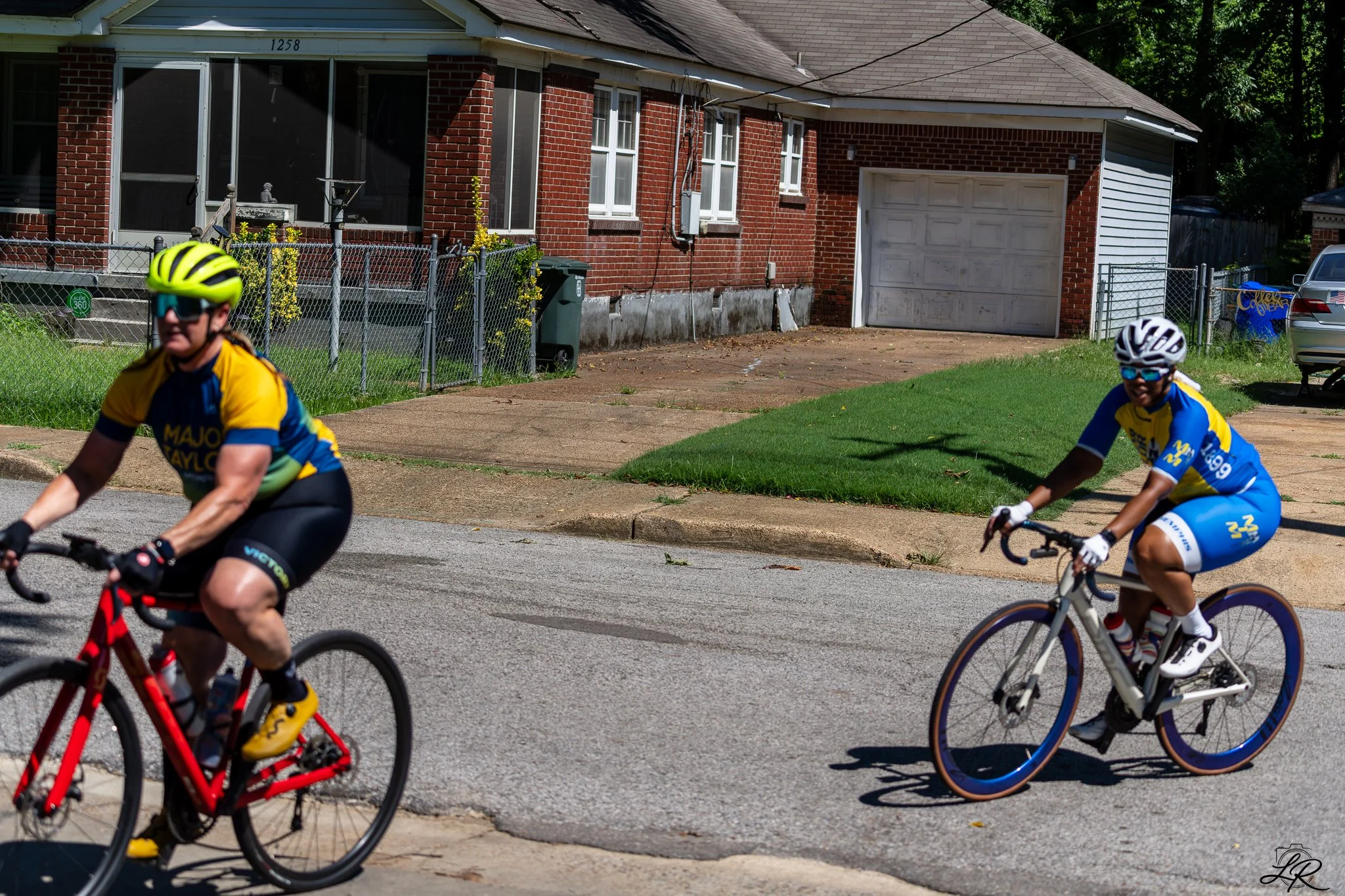 Two cyclists wearing yellow and blue jerseys riding on a street in front of a brick house with a white garage door, green lawn, and a chain-link fence.