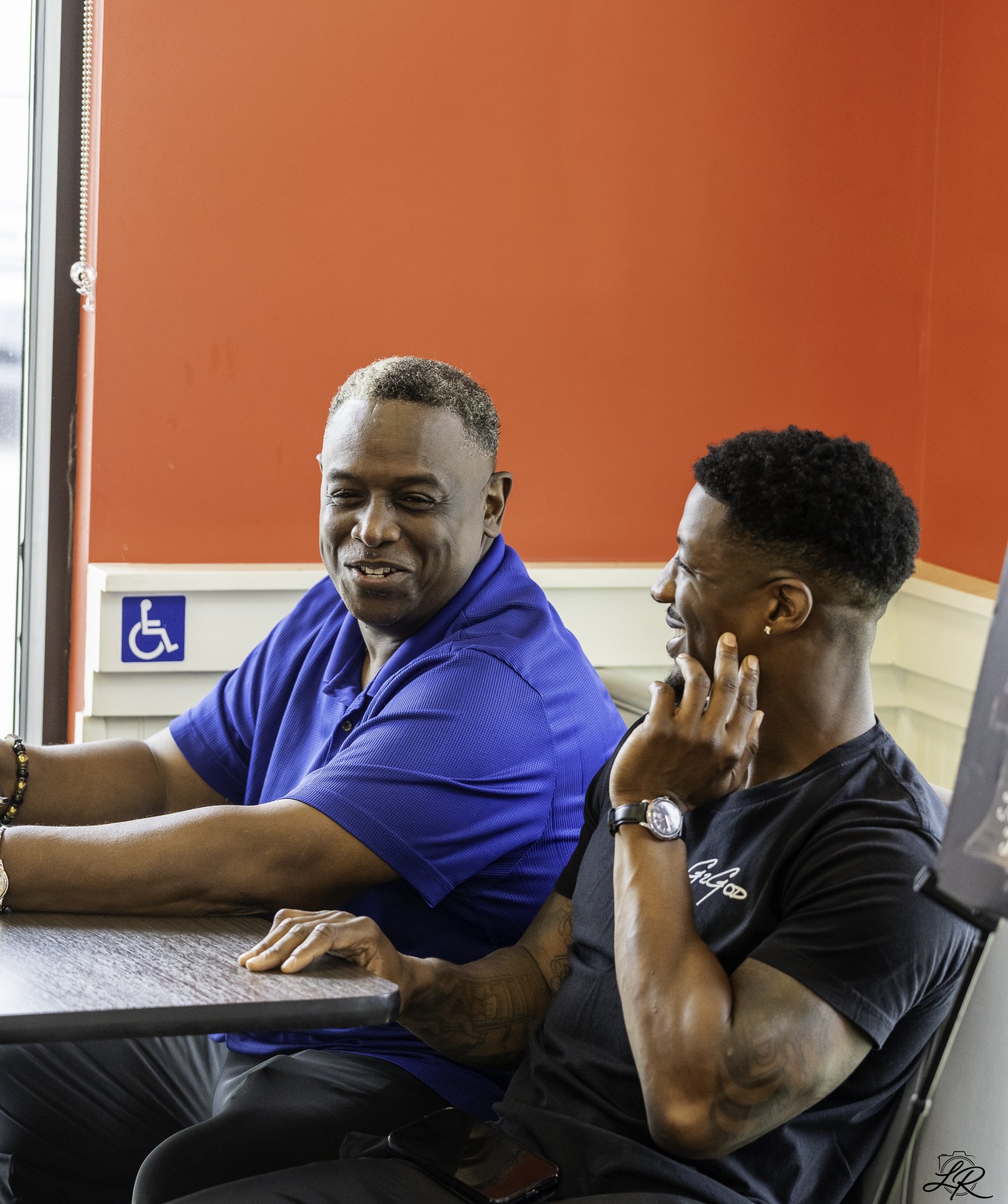 Two men sitting at a table, engaged in conversation. One man with a short curly hairstyle, wearing a blue shirt and glasses, smiling and leaning back slightly. The other man with short, styled hair, wearing a black shirt, touching his chin with his h