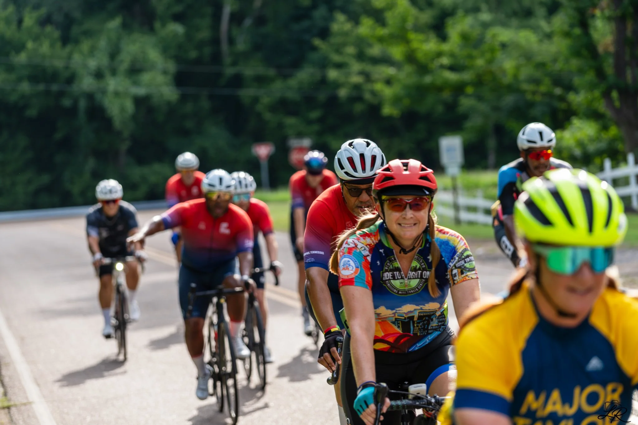 Group of cyclists riding on a scenic road surrounded by green trees, wearing helmets and colorful jerseys.