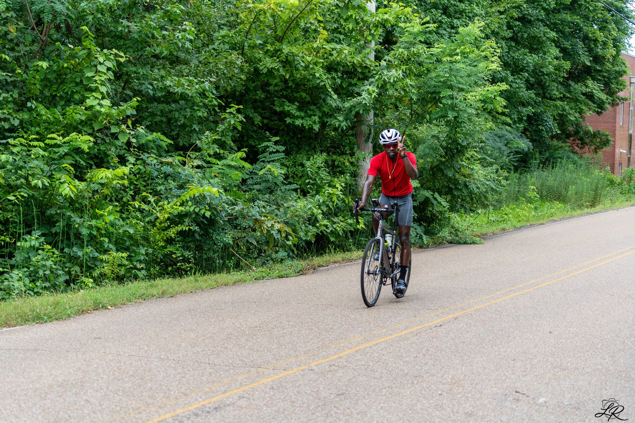 A man wearing a red shirt, gray shorts, and a white helmet riding a bicycle on a paved path next to green trees and shrubs.