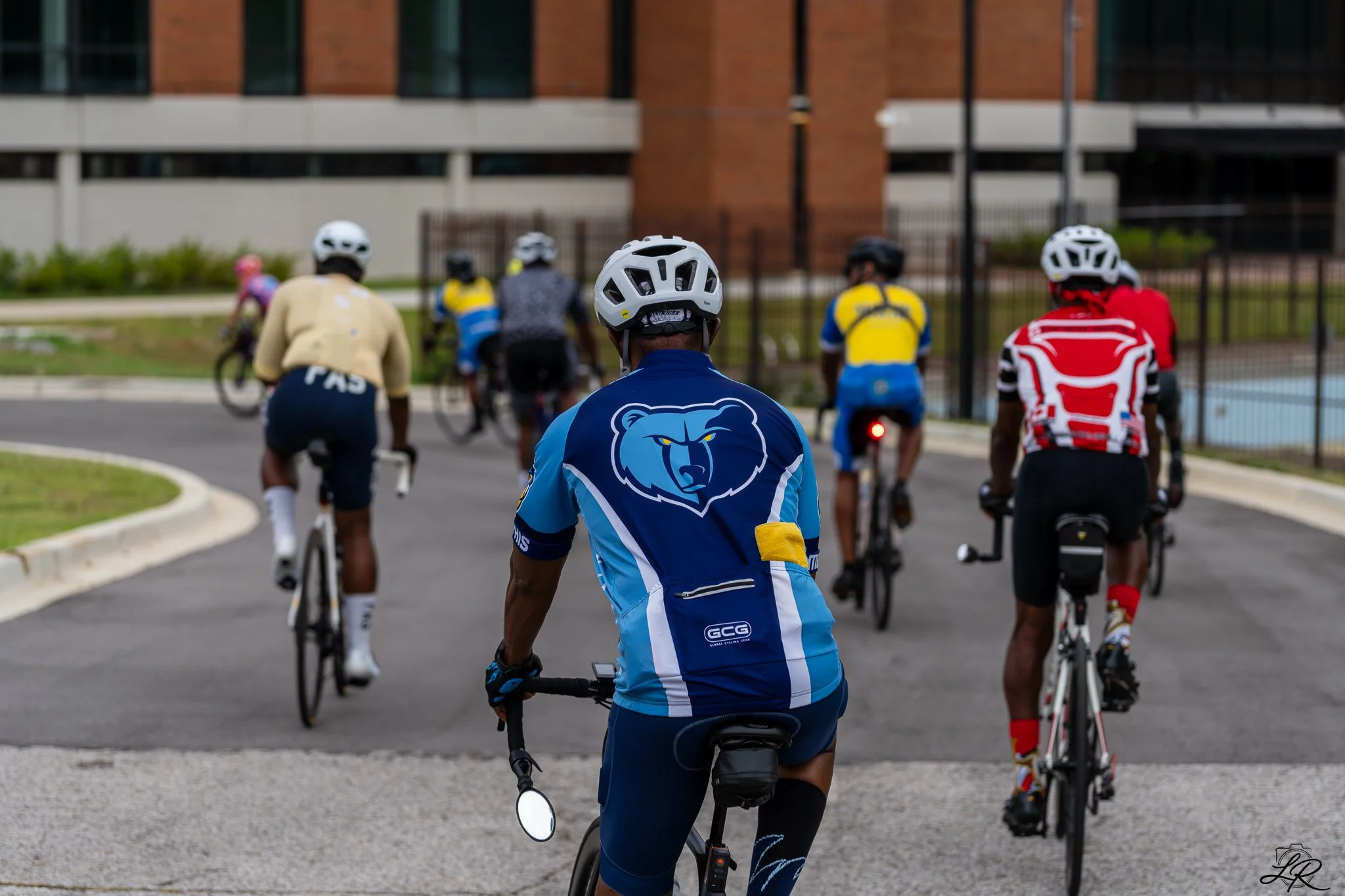 A group of people riding bicycles in an urban area, wearing helmets and athletic clothing, with a brick building in the background.
