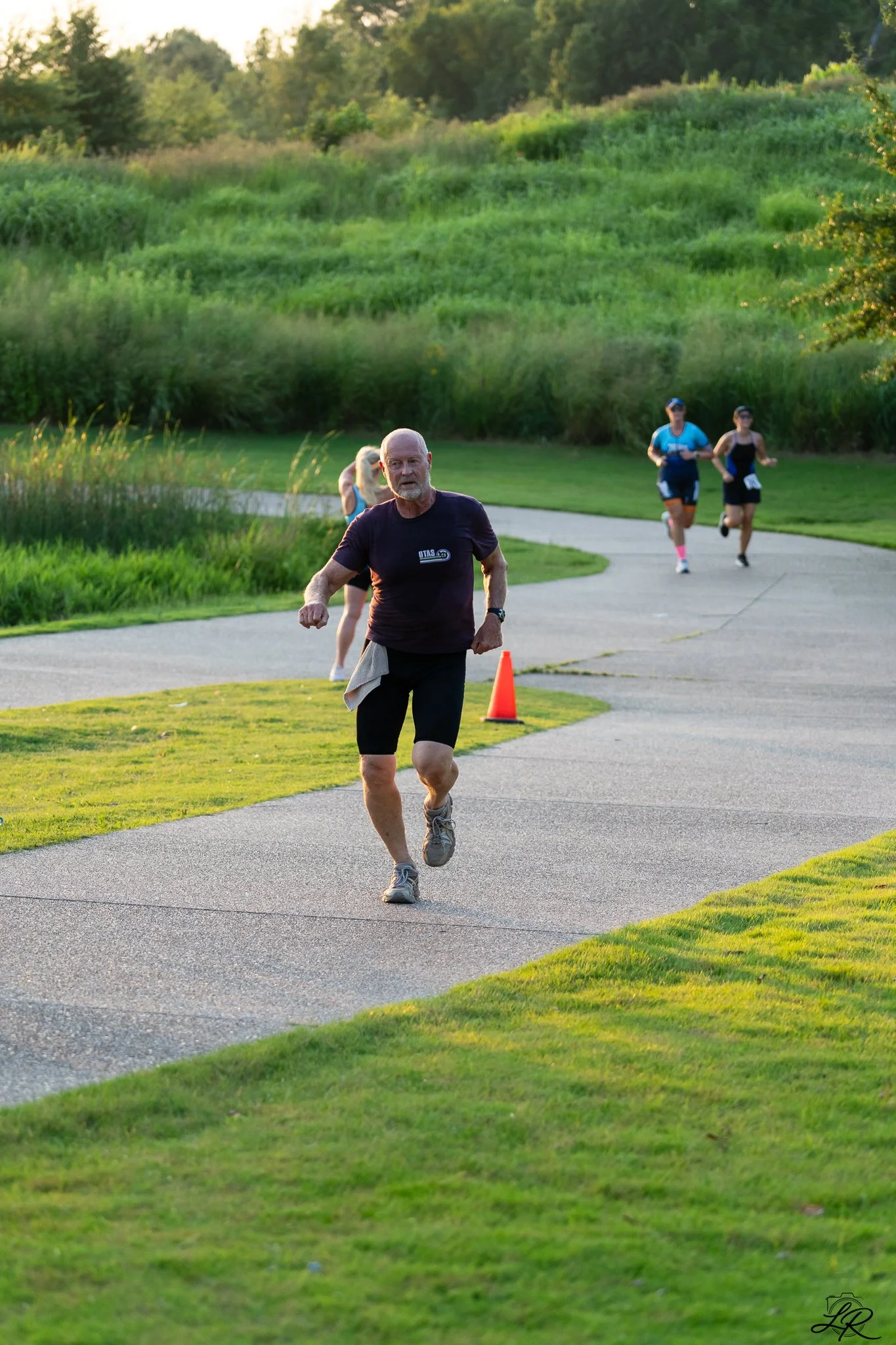 A man jogging on a curved paved path in a park during sunset, with a grassy field and trees in the background, accompanied by two women jogging behind him.