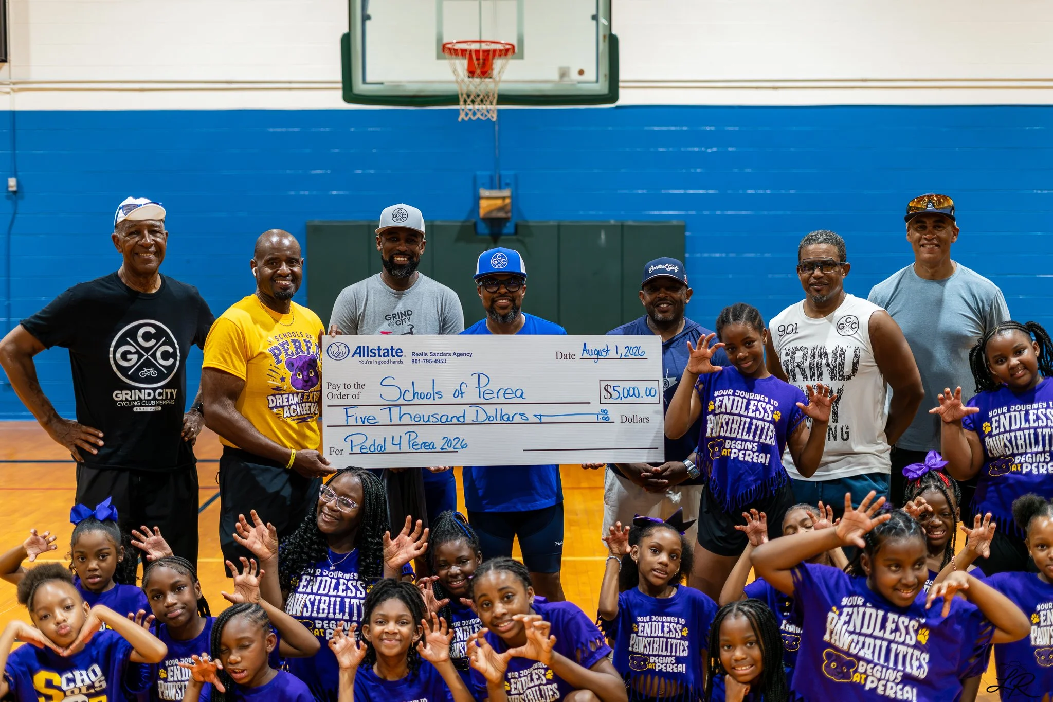 Group of adults and children in a gymnasium holding a large check for $5,000 made out to Schools of Perea for Pedal 4 Perea 2026, people smiling and posing for the photo.