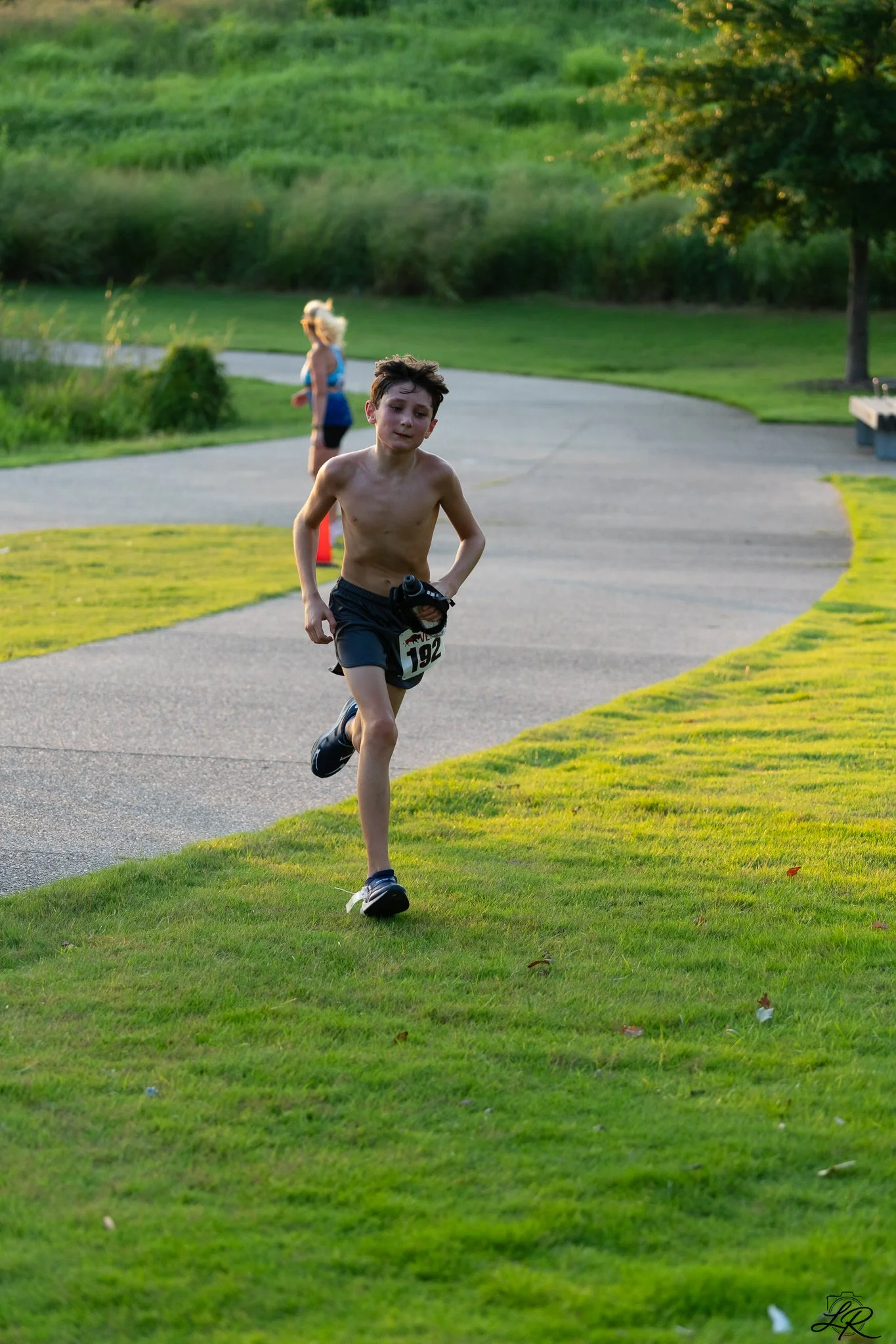 A young boy running outdoors on a grassy area near a paved path, with two women jogging in the background and trees and green grass surrounding them during sunset.