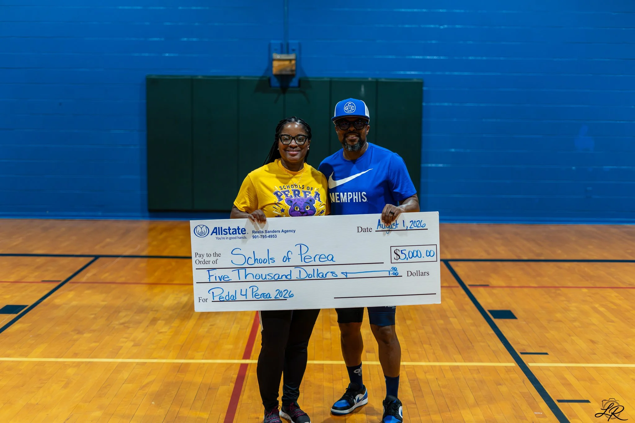 Two people inside a gymnasium holding a large check for five thousand dollars, dated August 1, 2026, made out to Schools of Perea for Pedal 4 Perea 2026. The person on the left wears a yellow T-shirt with a purple creature logo, and the person on the