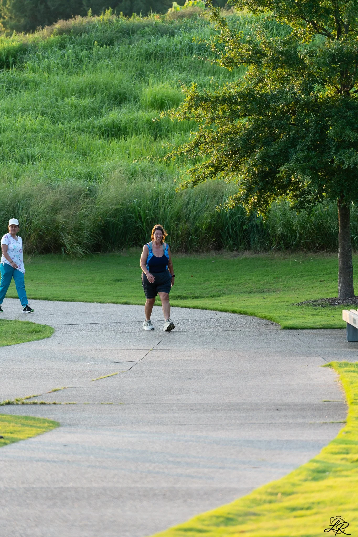 Two women walking along a curved concrete pathway in a park with green grass, trees, and a hillside covered in tall grass in the background, during late afternoon or early evening.