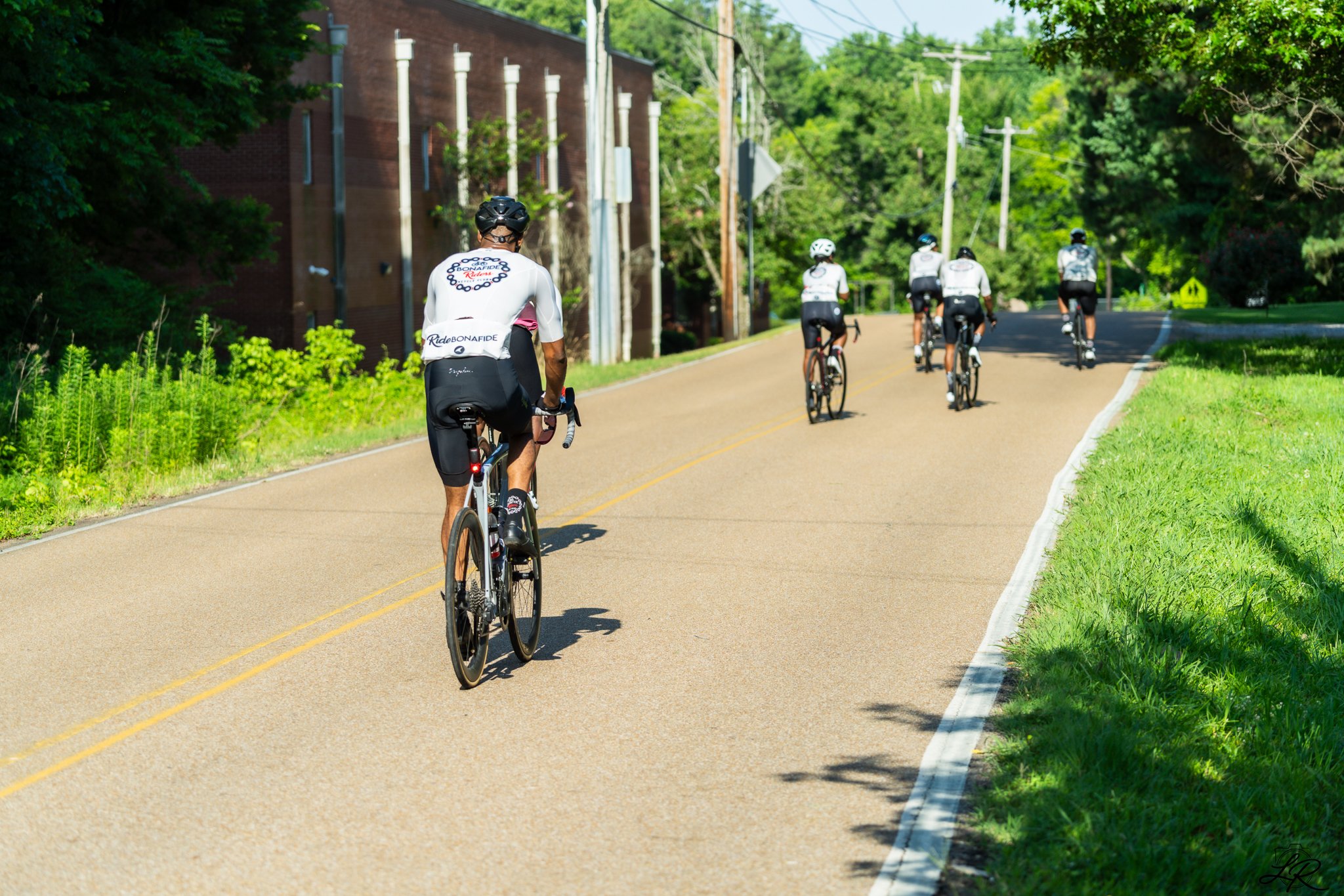 Group of five cyclists riding on a paved suburban road surrounded by greenery and trees, with utility poles and a brick building on the left.
