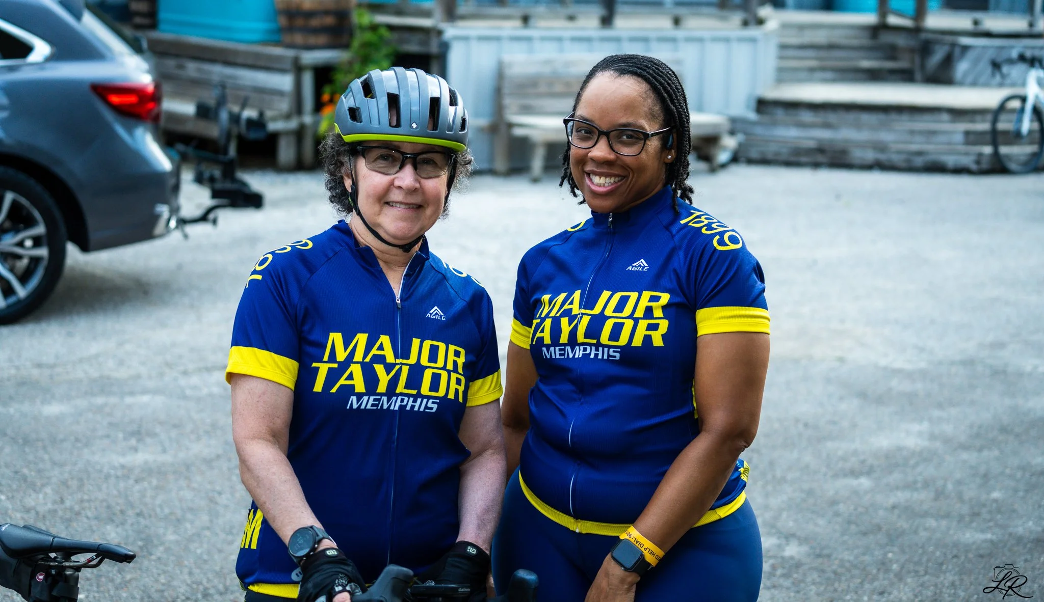 Two women in blue cycling jerseys with yellow accents and 'Major Taylor Memphis' written on them, smiling outdoors. One woman wears a helmet and glasses, the other wears glasses and has dreadlocks.