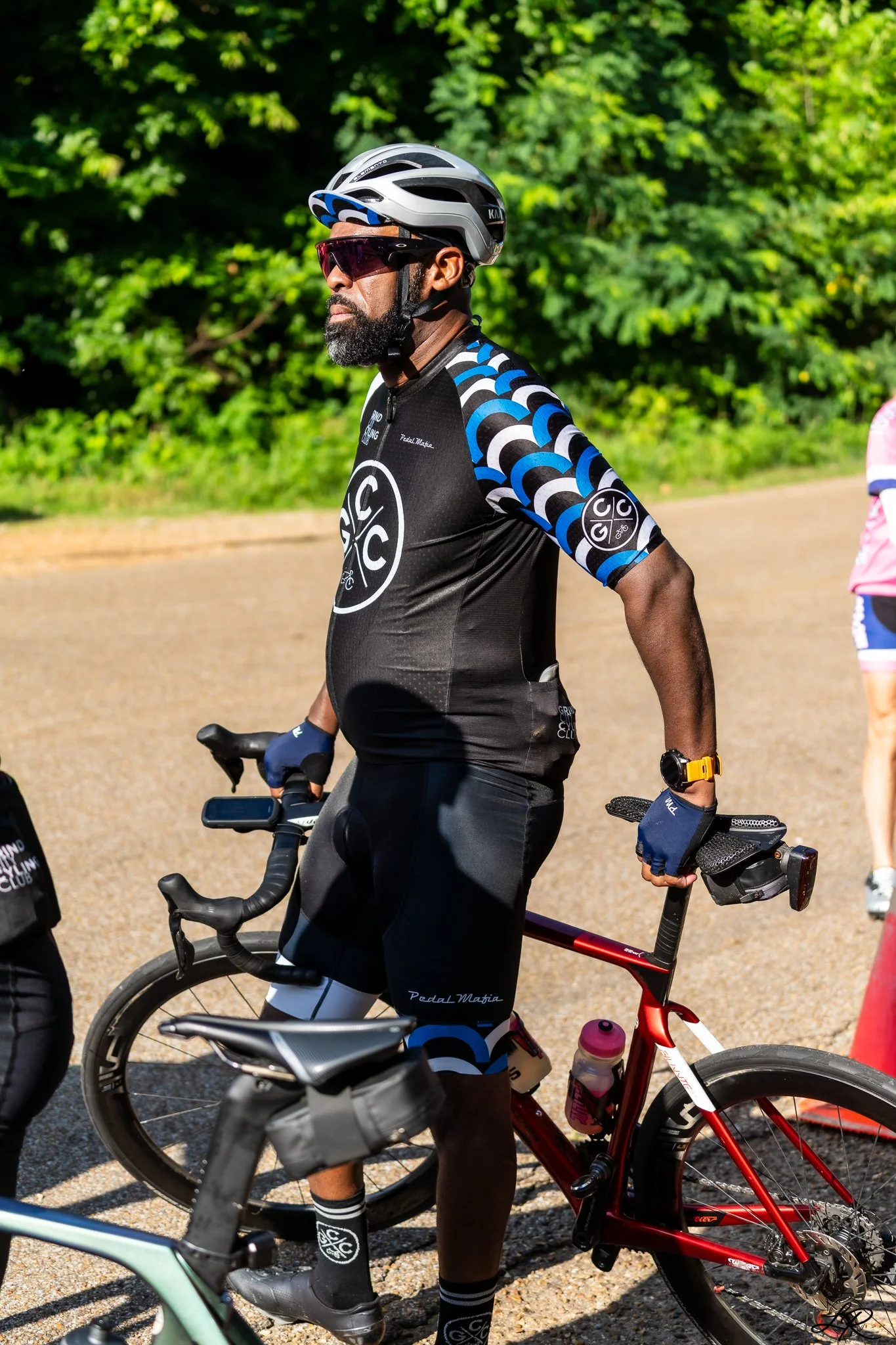 A man wearing a black cycling jersey with blue, white, and black patterned sleeves, black cycling shorts, a gray helmet, and tinted sunglasses, riding a red mountain bike on a gravel path with green trees in the background.