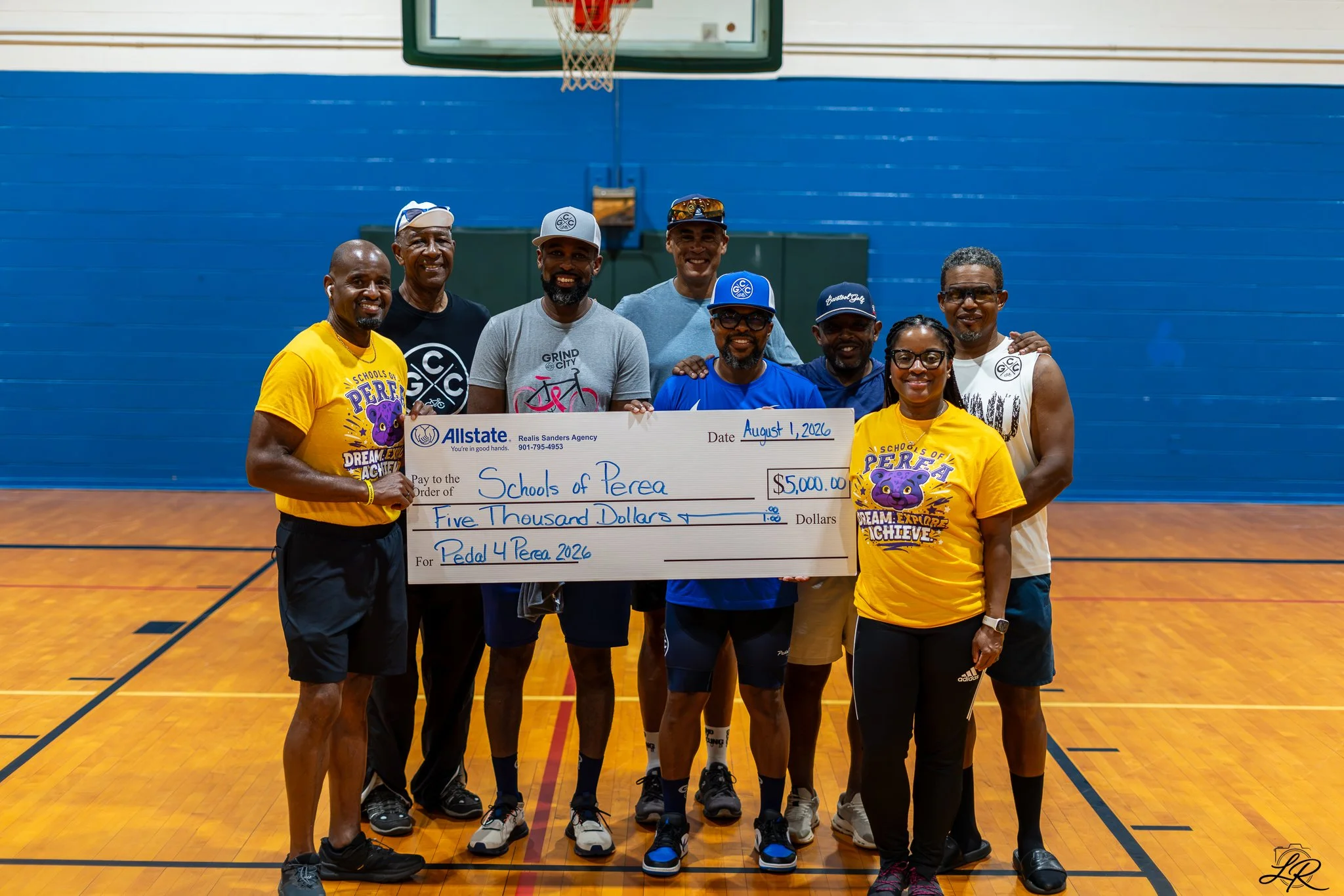 A group of nine people standing on a basketball court holding a large check for five thousand dollars, dated August 1, 2026, made out to 'Schools of Perea' for 'Pedal 4 Perea 2026.' They are smiling and wearing casual athletic clothes, some with caps