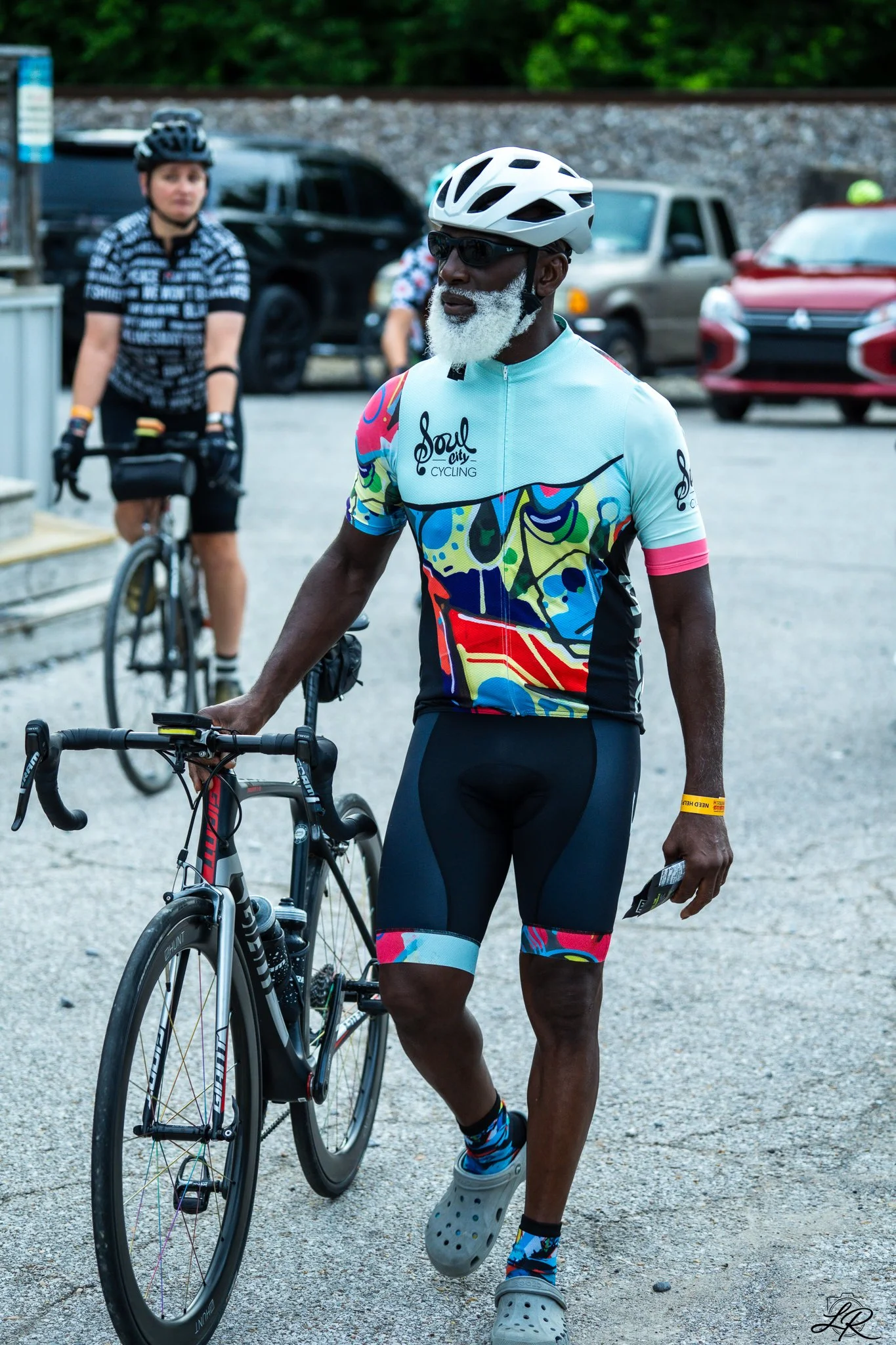 A man with a white beard, wearing a colorful cycling jersey, black shorts, a white helmet, sunglasses, and white Crocs, stands next to a black road bicycle. Other cyclists and parked cars are visible in the background.