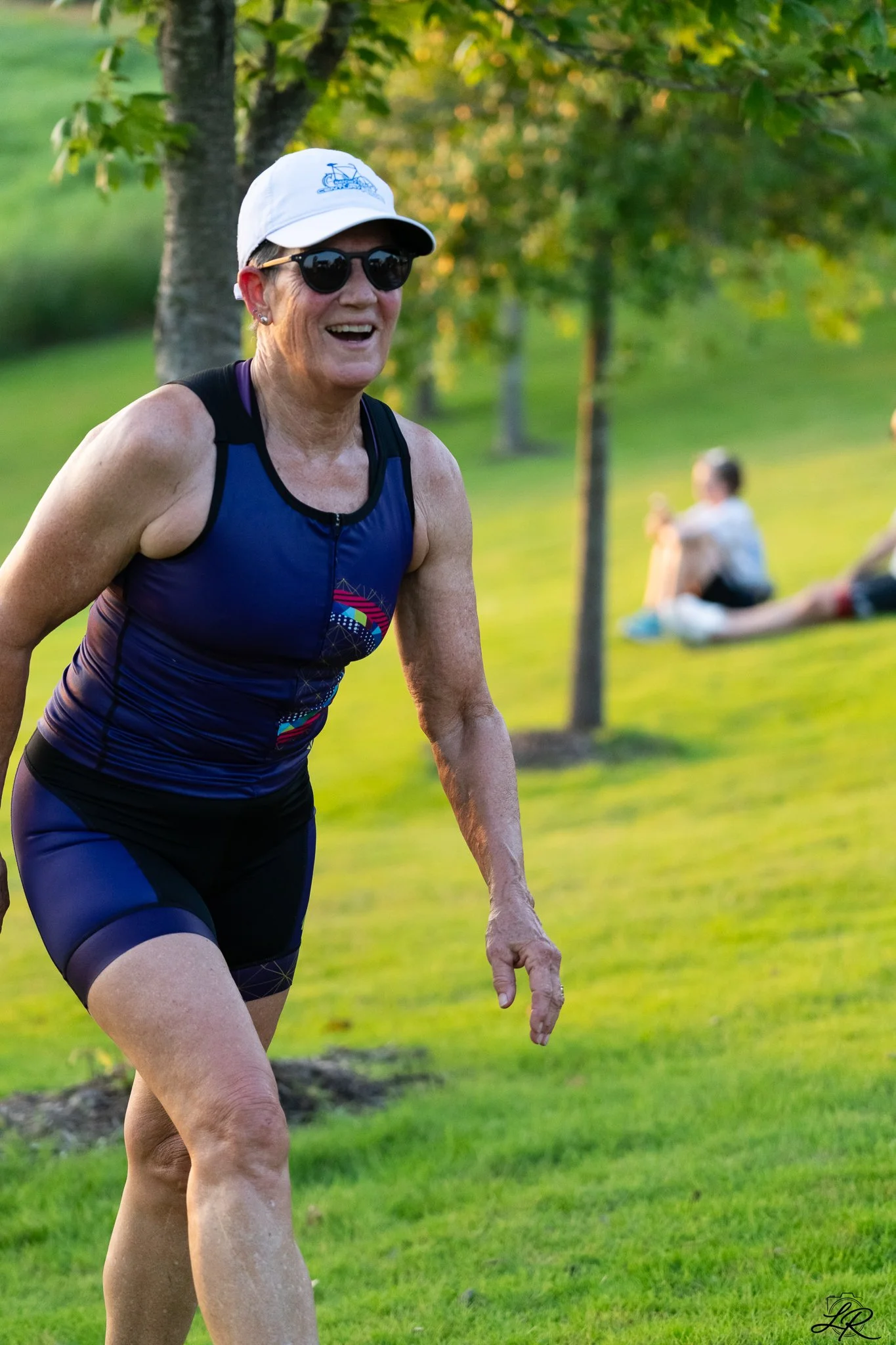 A smiling older woman wearing sunglasses, a white cap, and athletic clothing participates in an outdoor running event in a park with green grass and trees, with other people sitting on the grass in the background.