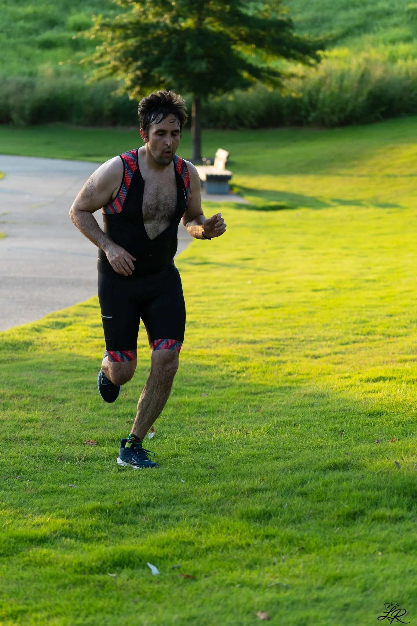 A man running on a grassy park with trees and a bench in the background during late afternoon.