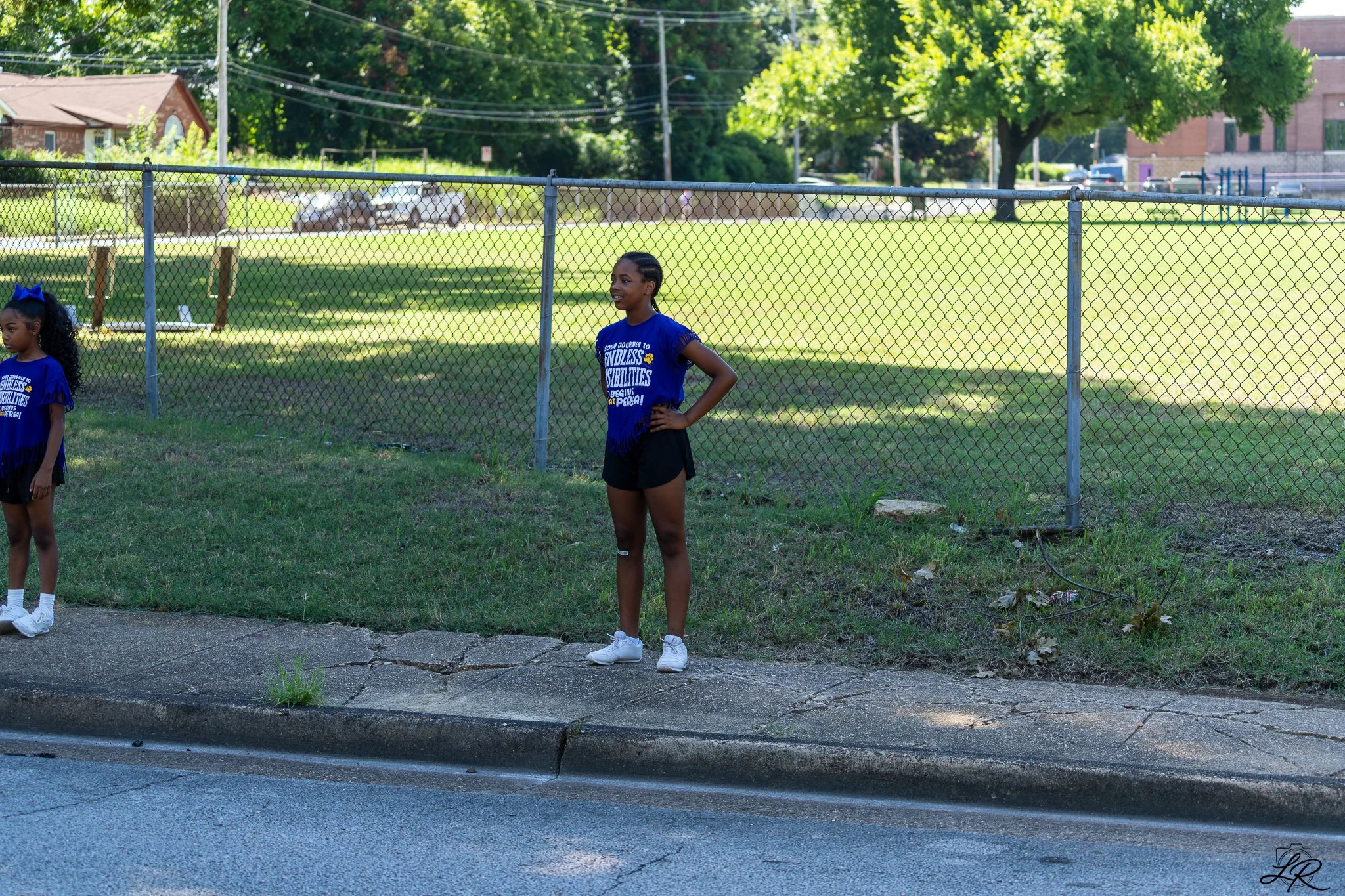 Two young girls standing outdoors near a chain-link fence on a sunny day. Both are wearing blue T-shirts and black shorts. The background features green trees, a grassy area, and some parked cars.