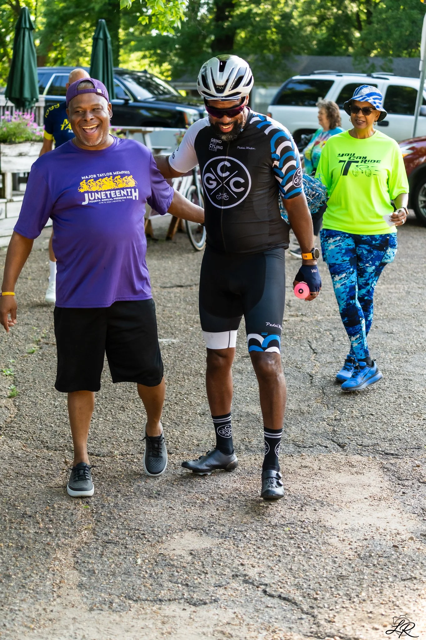 A group of people outdoors, including a man in sports gear with a bicycle logo on his shirt, walking with a man in a purple shirt that reads 'Major Taylor Memphis Unofficial'. A woman in bright yellow sportswear and a hat follows behind. In the backg