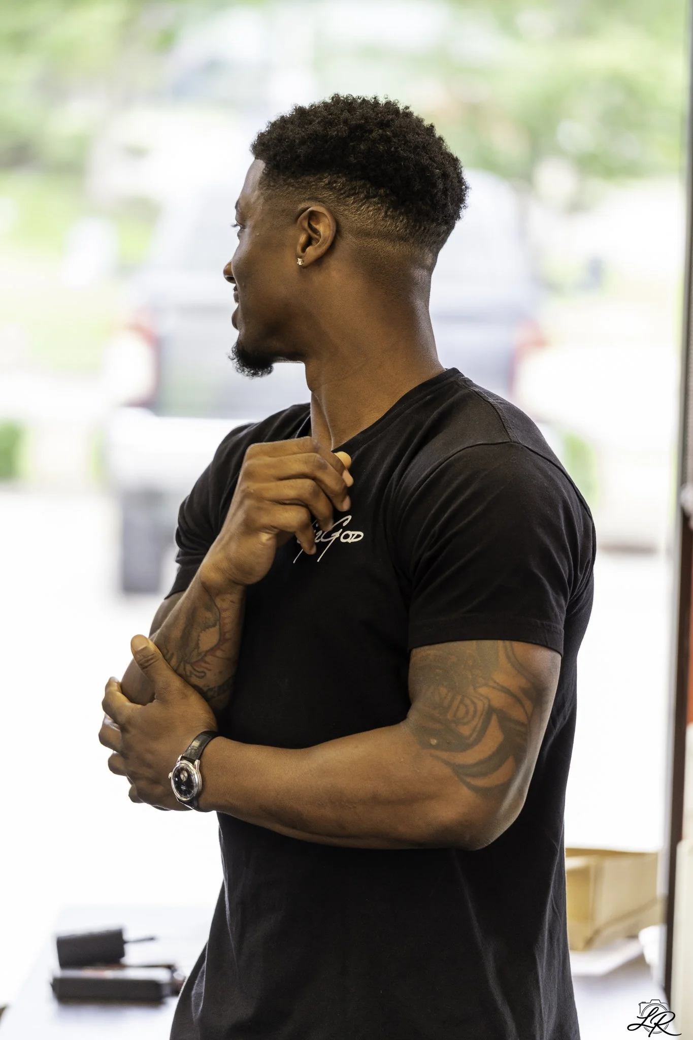 Profile of a young man with curly hair and tattoos, wearing a black T-shirt, indoors near a window, adjusting his collar with a watch on his left wrist.