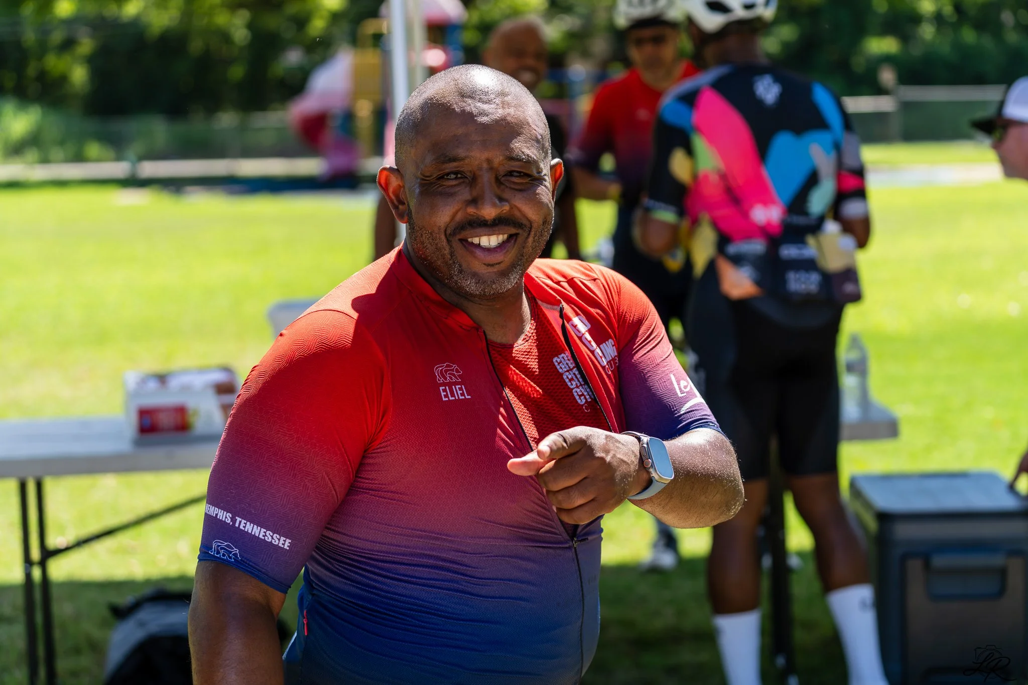 Smiling man in a red and blue athletic shirt with a smartwatch on his wrist, standing outdoors at a sporting event, with a blurred background of people and greenery.