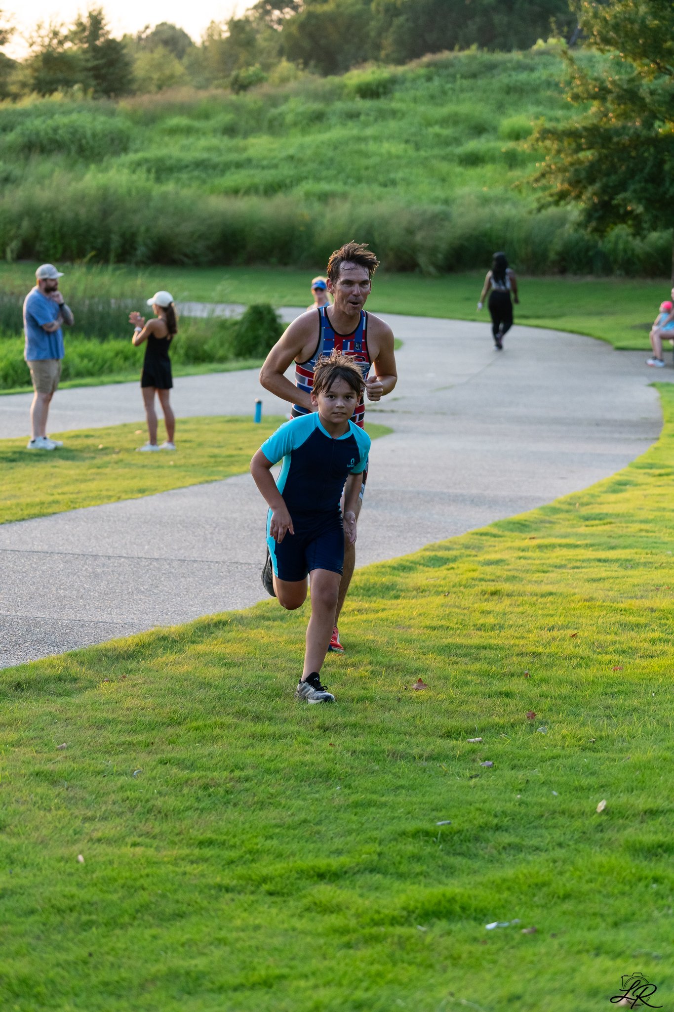 A man and a boy running on a grassy path in a park, with several people standing and walking in the background, surrounded by green trees and hills.