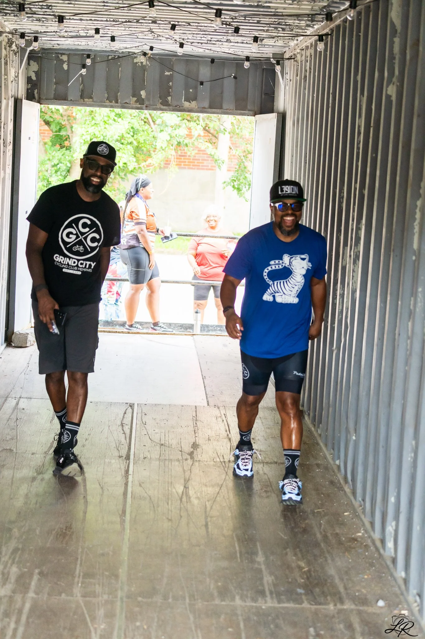 Two men walking through a metal-framed tunnel, with one wearing a black t-shirt with white graphics and shorts, and the other in a blue t-shirt with a tiger graphic and shorts. Both are smiling, wearing sunglasses, hats, and running shoes. In the bac
