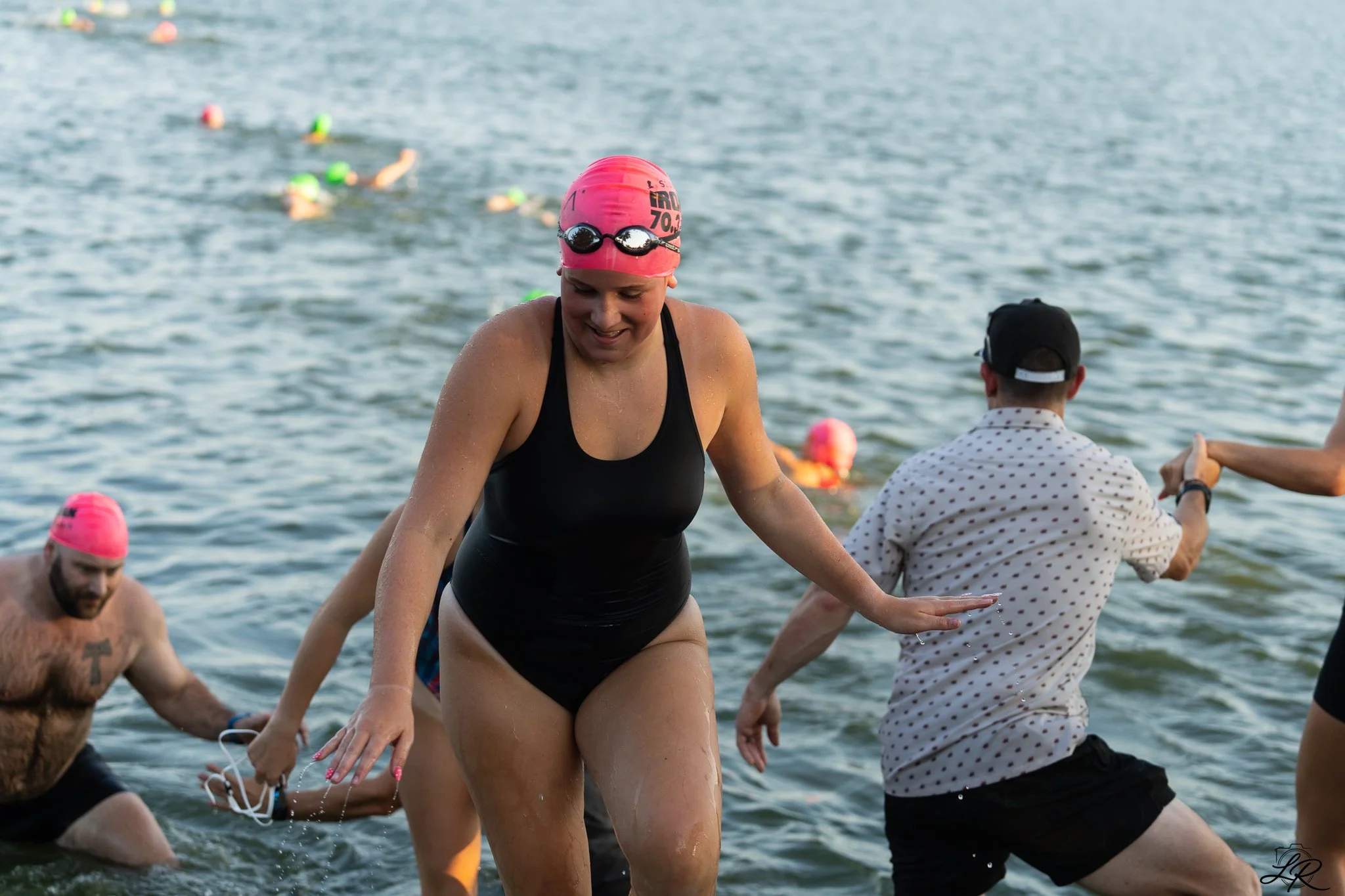 Swimmers wearing bright green and pink swim caps and goggles in a lake, with some helping each other out of the water.