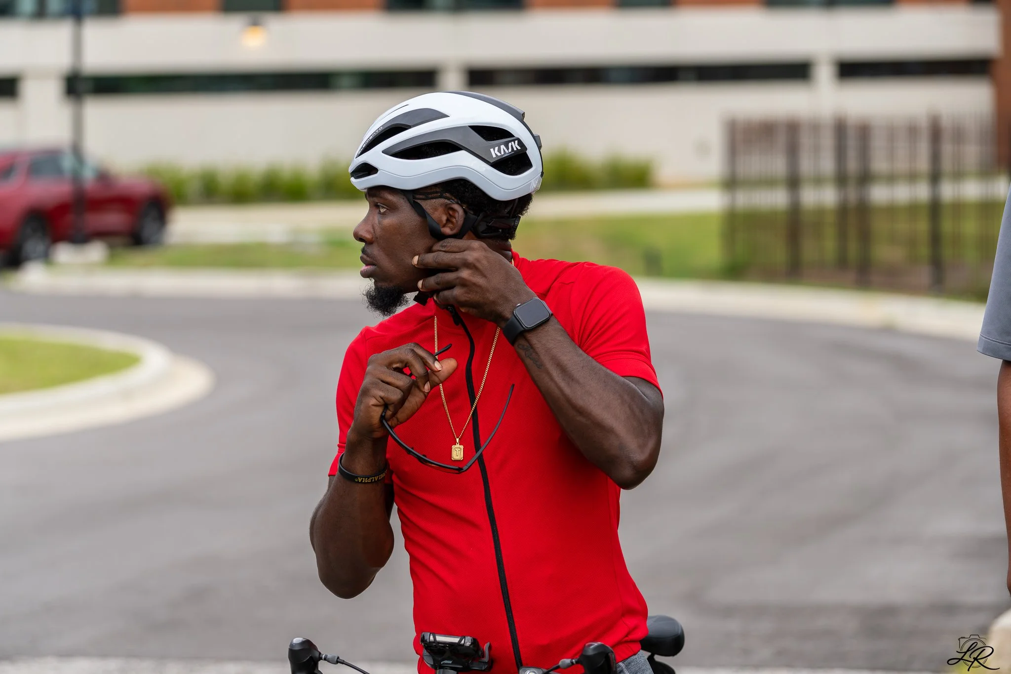 A man wearing a white bicycle helmet and a red shirt adjusts his helmet strap while standing next to a bicycle, outdoors in a parking lot with a blurred building and cars in the background.