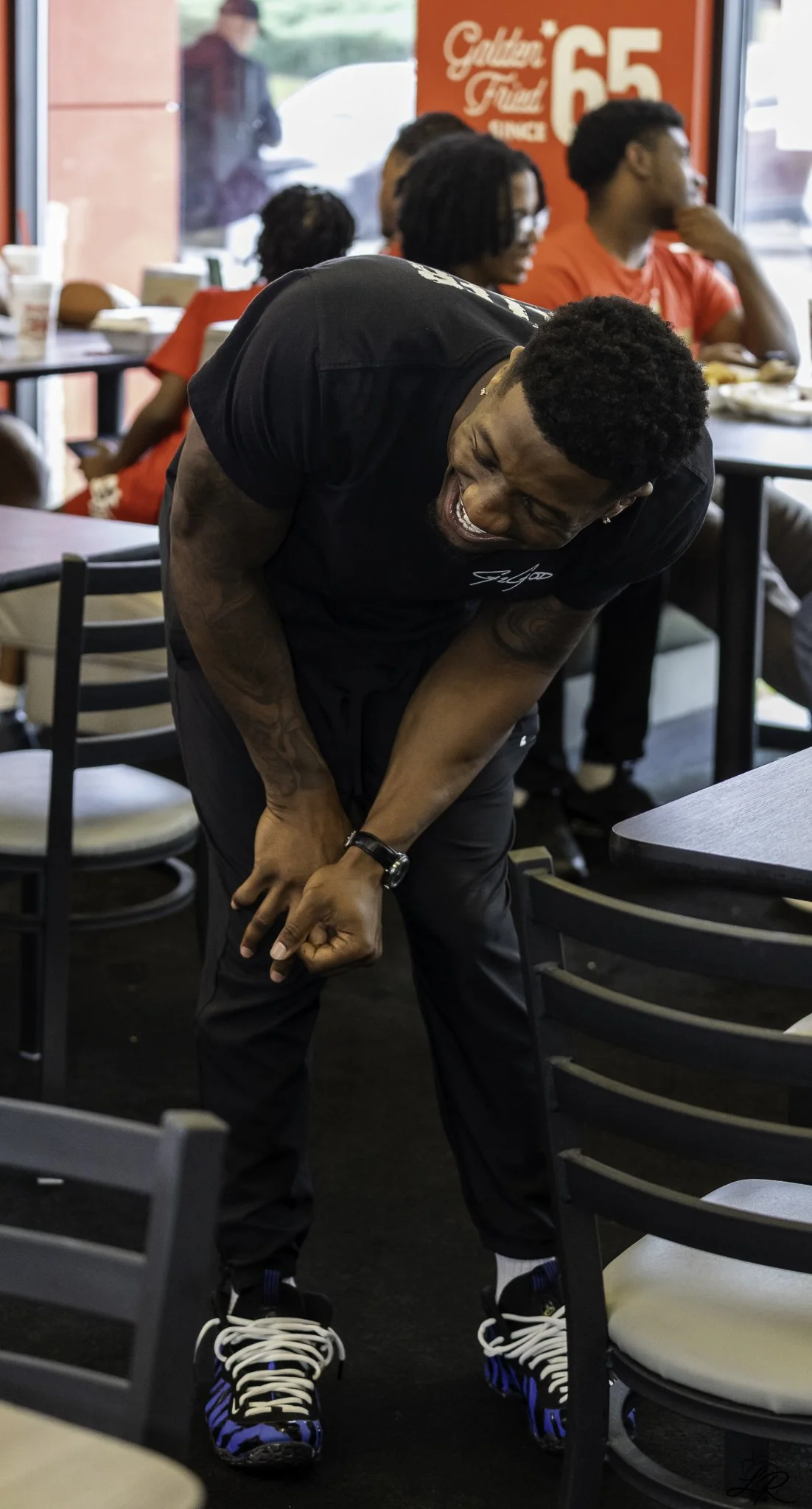 A young man with black curly hair, wearing a black t-shirt and black pants, is bending over and laughing inside a restaurant. He is wearing black and white sneakers with blue accents. In the background, there are several people sitting at tables, and