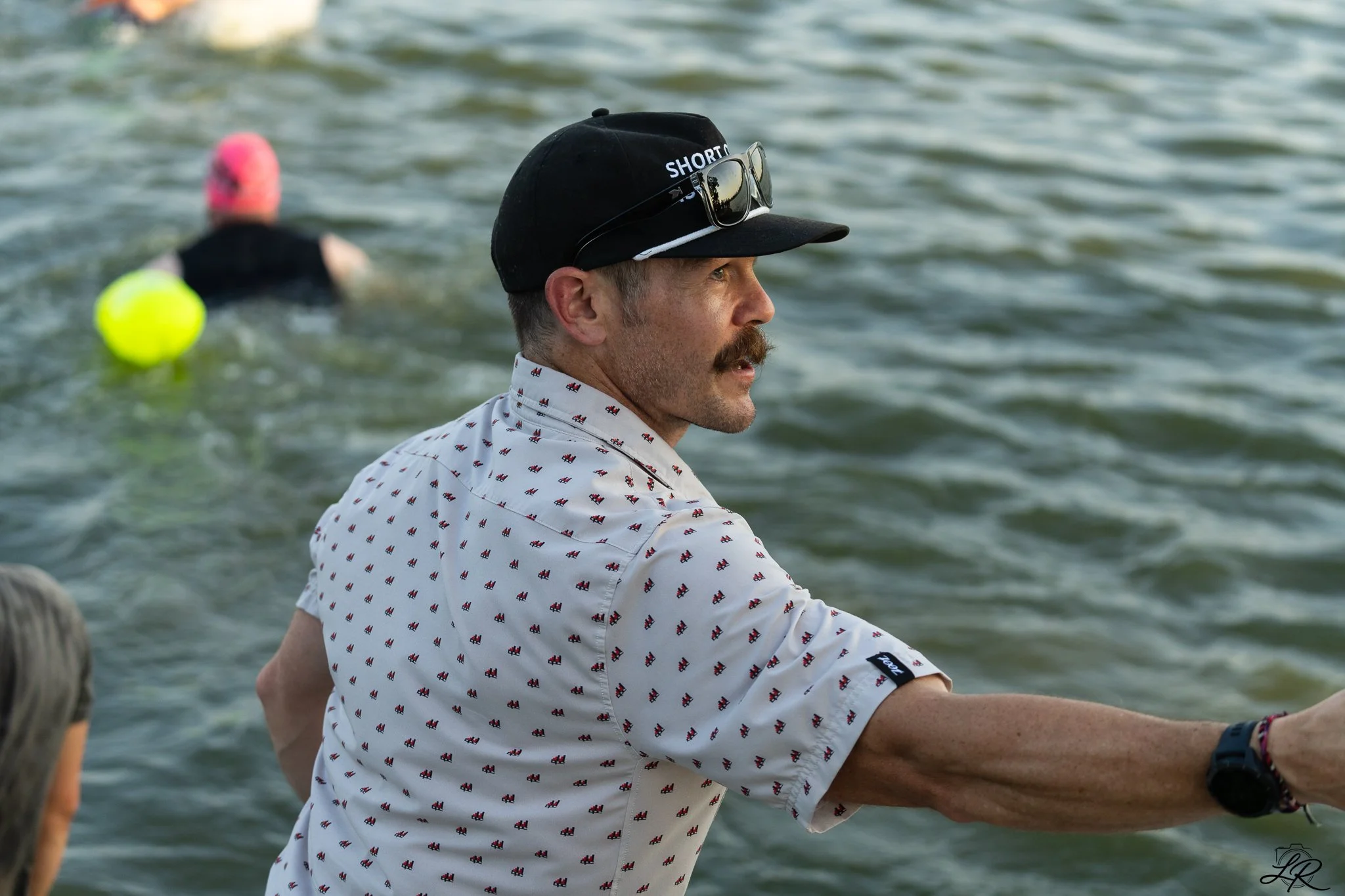 A man with a mustache wearing a white shirt with a small pattern, a black cap with sunglasses on top, sits on a boat or pier near water, with another person in the background wearing a pink swim cap and dark swimsuit, and a yellow ball in the water.