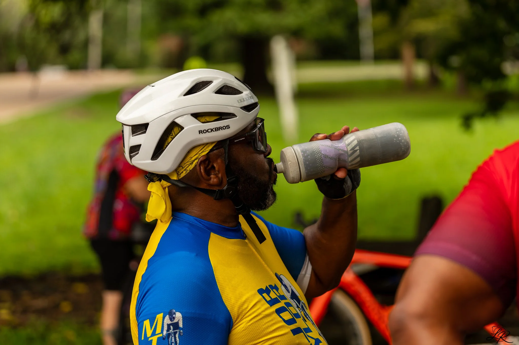 Close-up of a man wearing a white bike helmet and blue and yellow cycling jersey, drinking from a gray water bottle outdoors with blurred trees and people in the background.