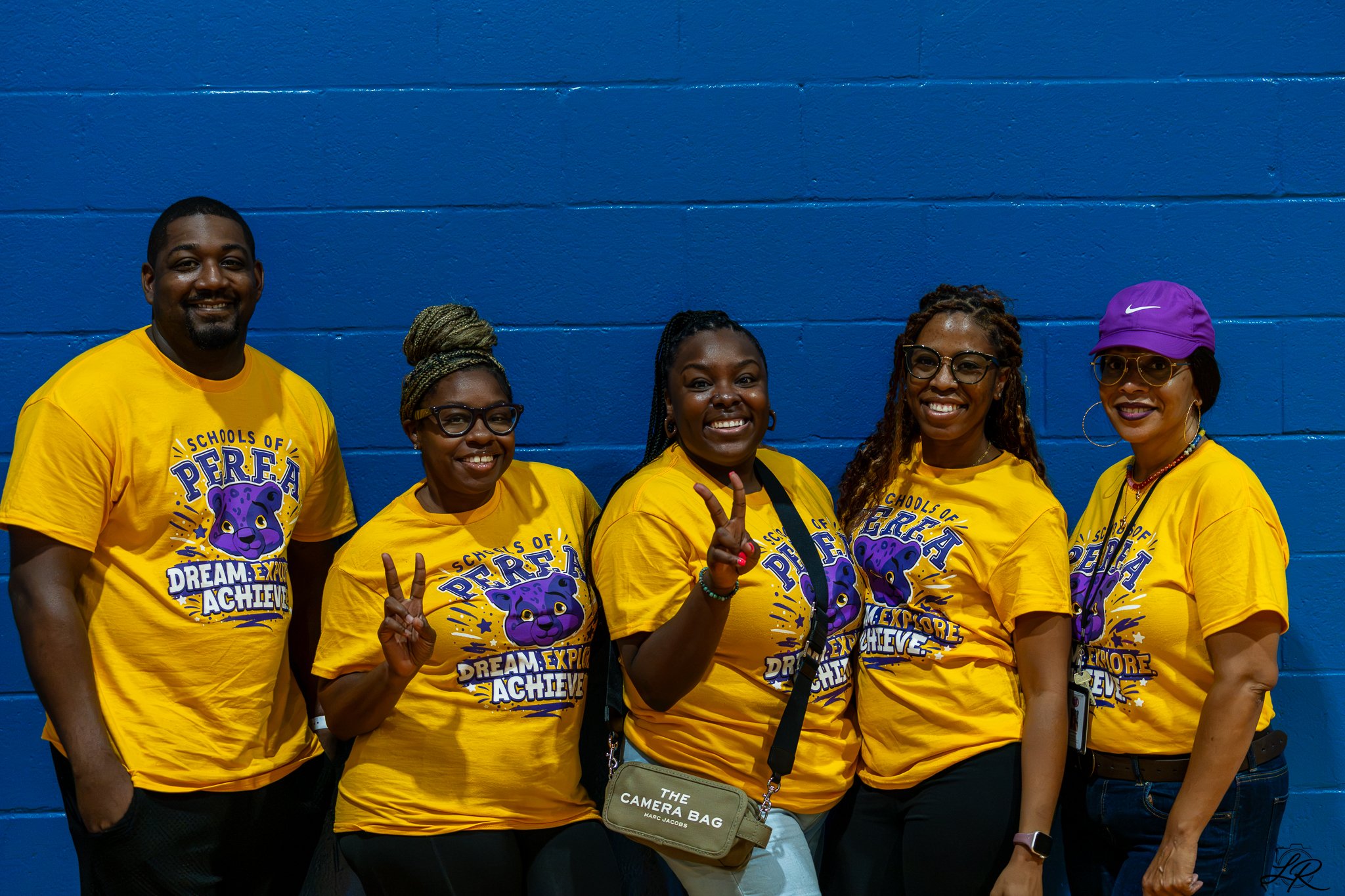 Group of five diverse adults wearing matching yellow T-shirts with purple bear graphic and motivational text, standing against a blue wall, smiling and making peace signs.