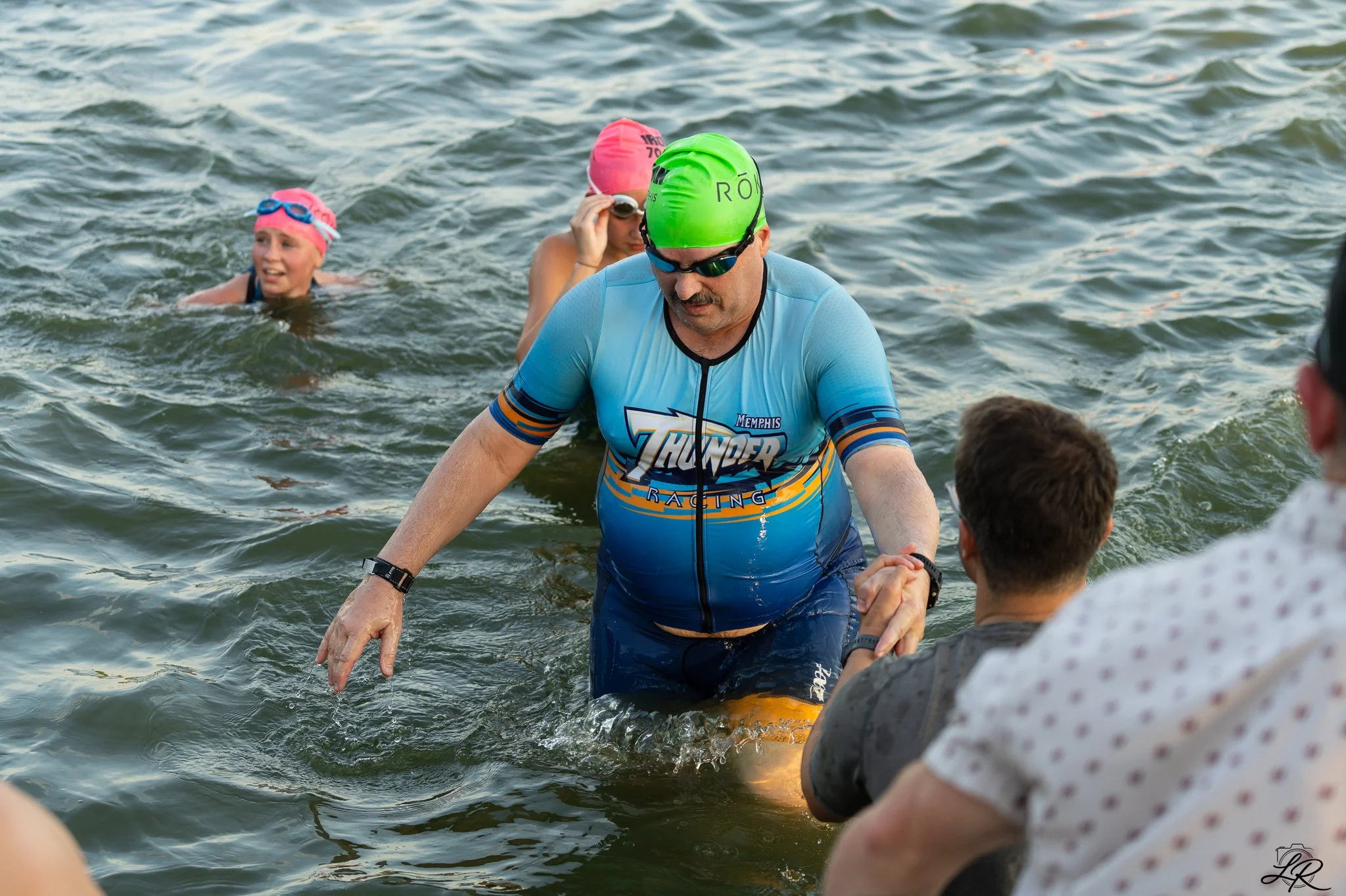 A man in a blue cycling jersey and green helmet in the water, shaking hands with a person outside of the water. Two women wearing pink swim caps and goggles are swimming nearby in the water.