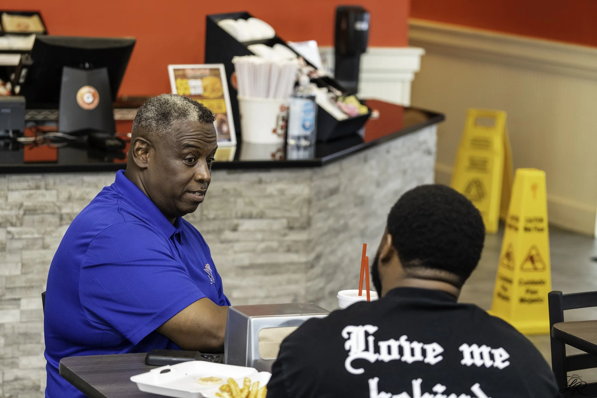 A man in a blue shirt sits at a table inside a fast-food restaurant, talking with a young boy with short hair wearing a black shirt with white text. The restaurant has red walls, a cash register, and a yellow caution sign on the floor.