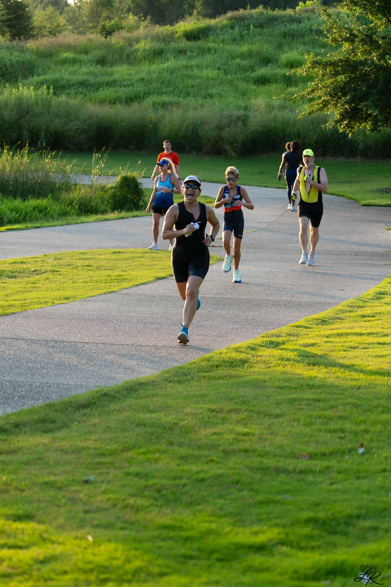 Group of seven runners jogging on a paved trail in a lush green park during late afternoon or early evening.