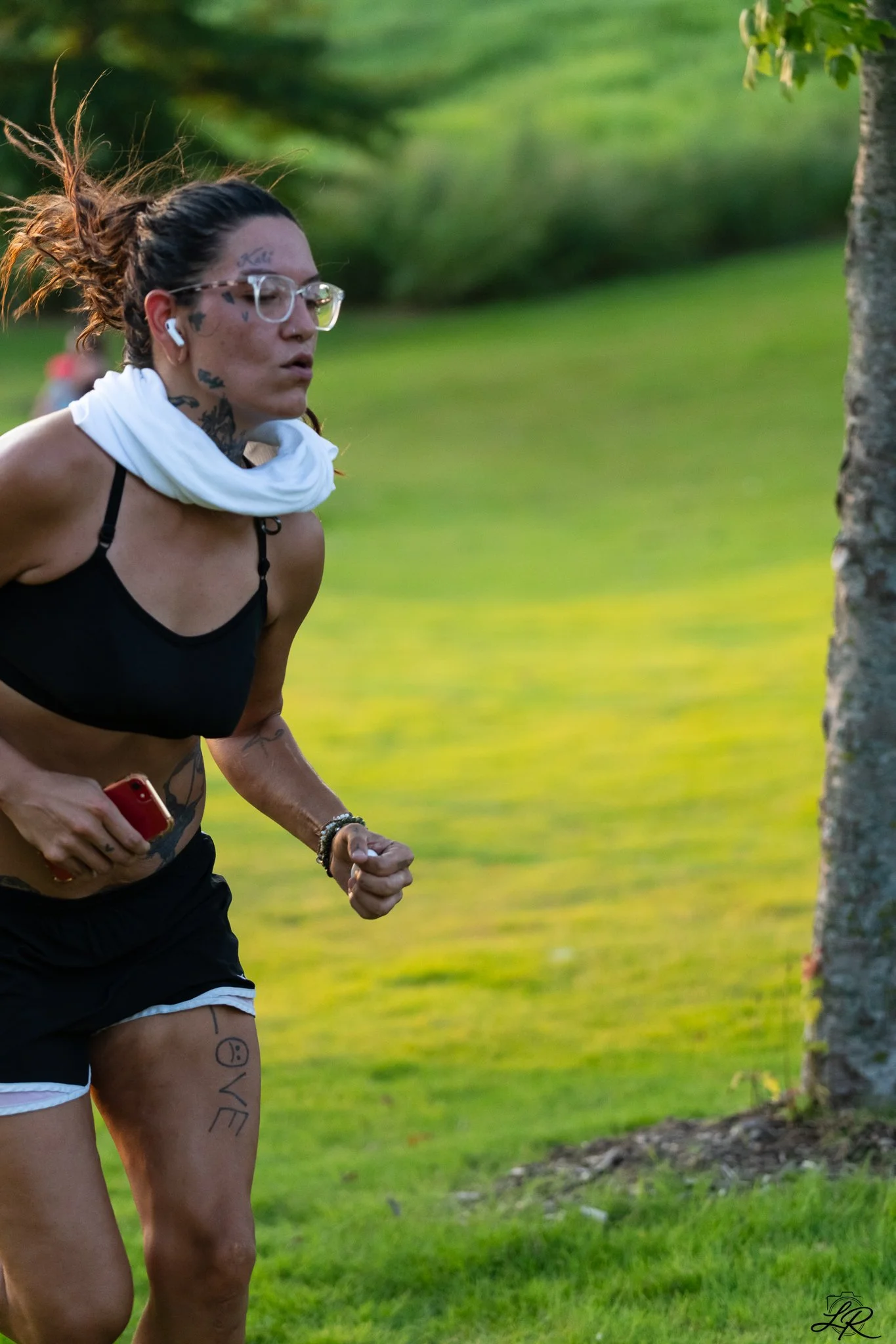 A woman running outdoors on a grassy area, wearing glasses, a black sports bra, black shorts with white trim, and AirPods, with clock tattoos and the word LOVE tattooed on her thigh.