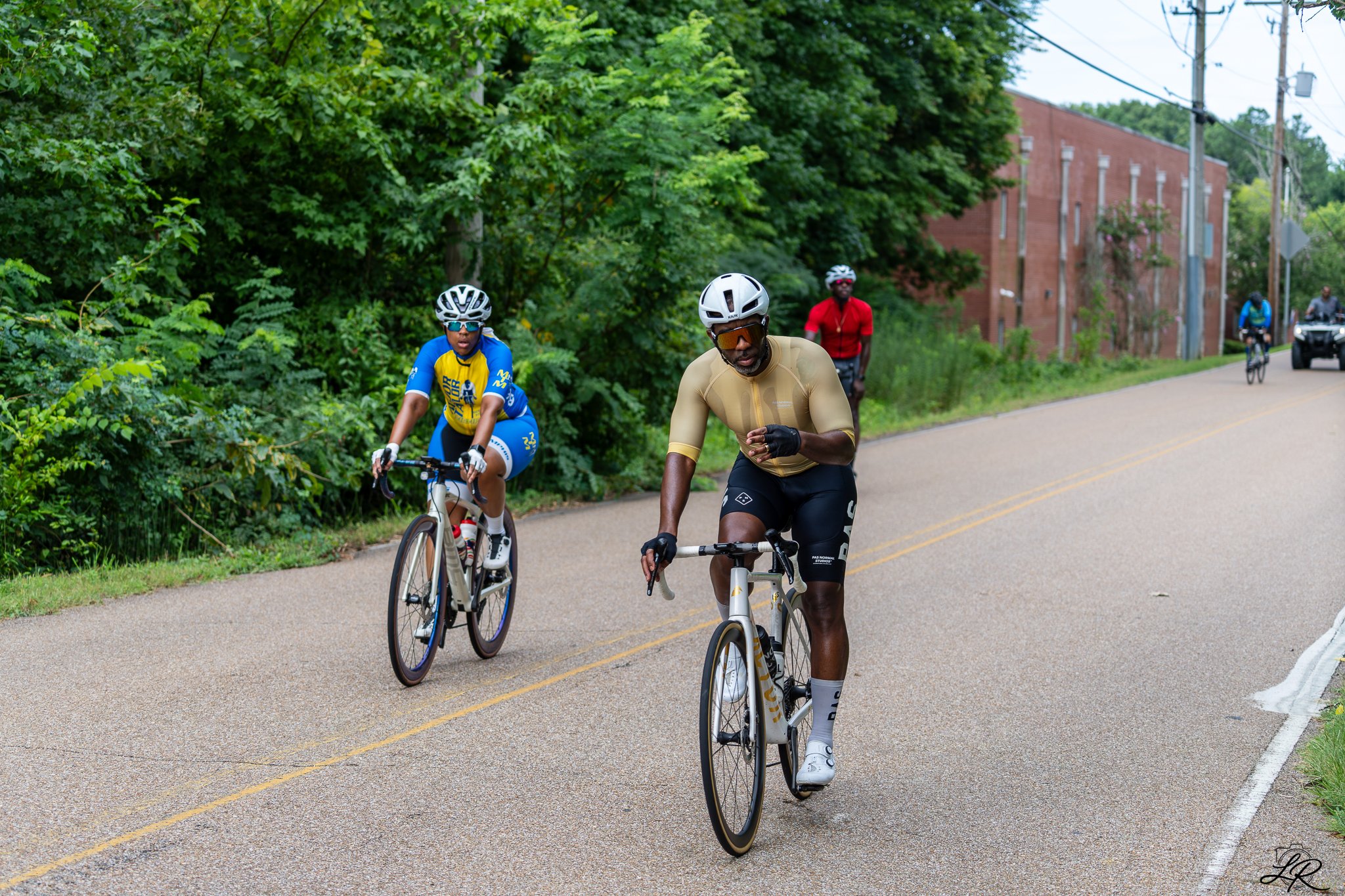 A group of cyclists riding on a rural road with greenery on one side and a brick building on the other, wearing helmets and sportswear.