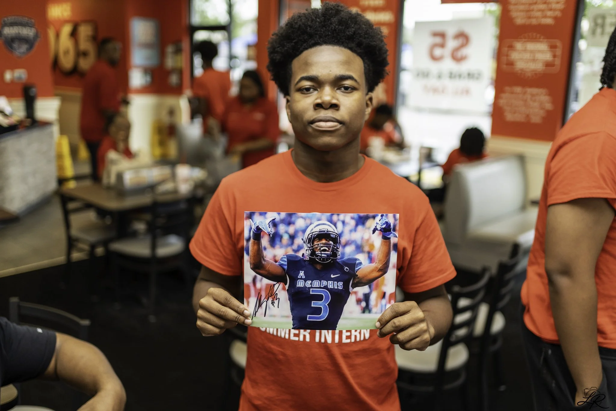 A young man with curly hair wearing a red T-shirt holding a signed photo of a football player in a blue jersey with the word Memphis.