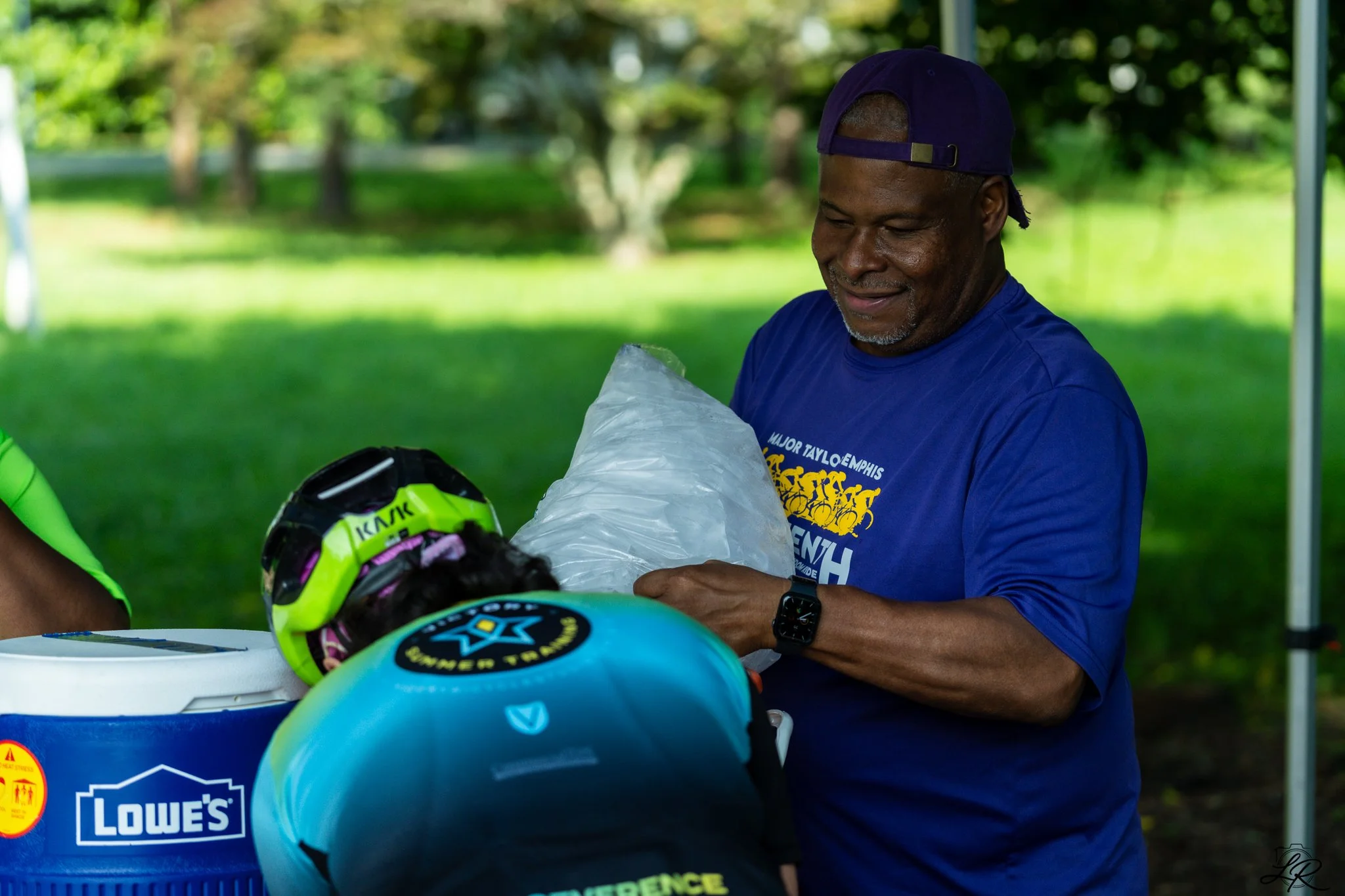 A smiling man in a purple cap and blue shirt stands outdoors, holding a plastic bag, with helmets on a table in front of him