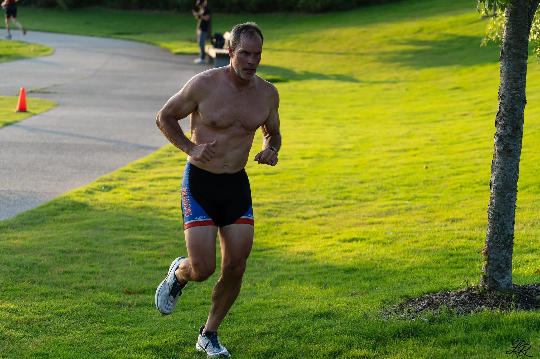 A shirtless man running outdoors on a grassy area, wearing black athletic shorts with red and blue accents and white running shoes.