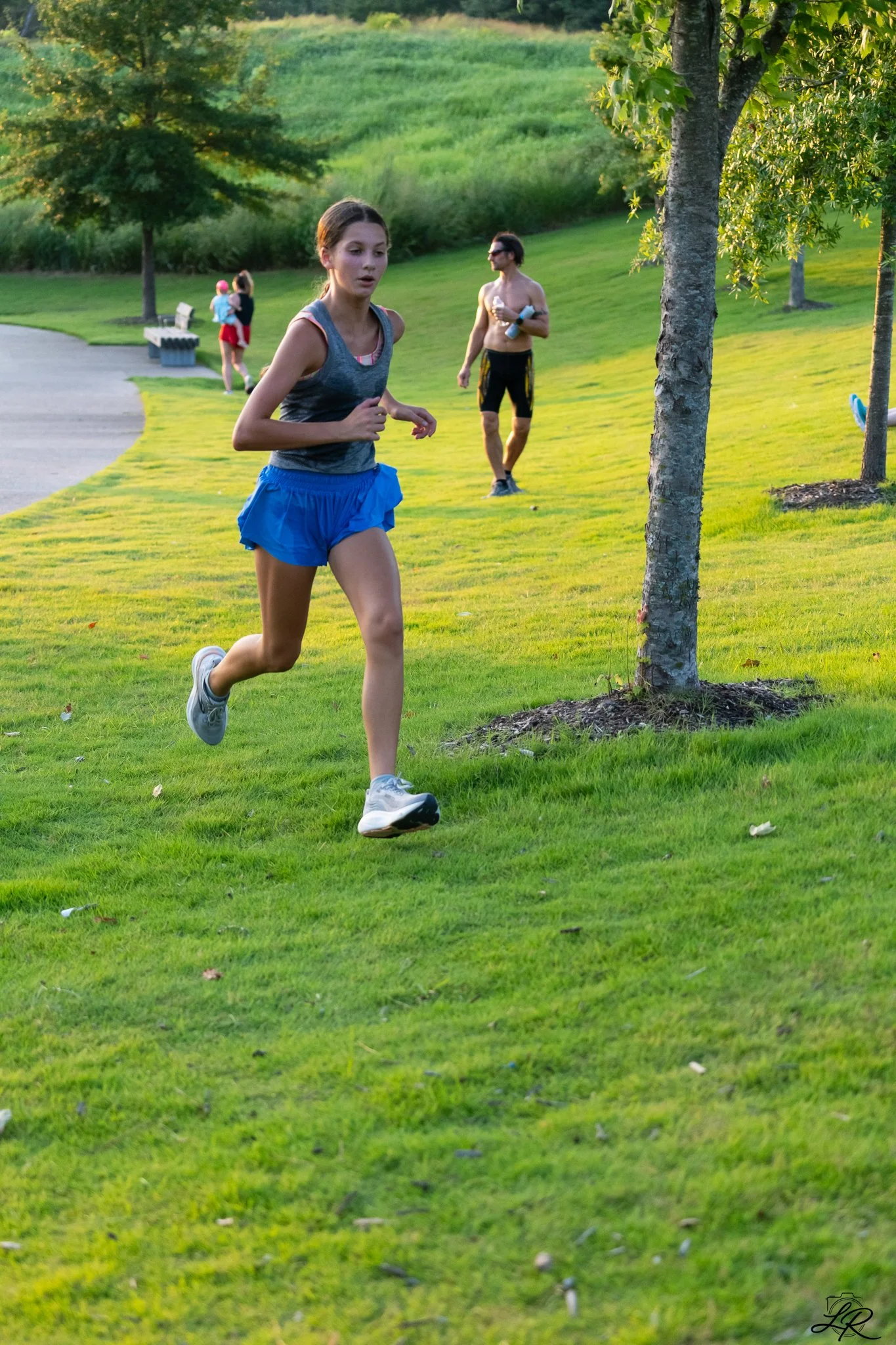 A young woman in athletic wear jogging on a grassy park path, with a shirtless man holding a water bottle walking behind her, and two women in the background near a bench and trees.