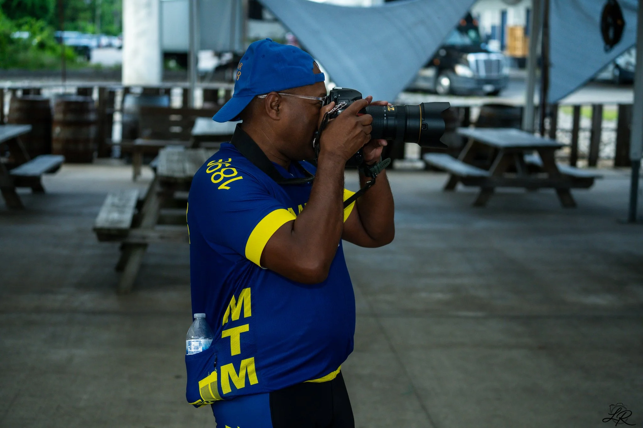 A person wearing a blue cap and a blue and yellow shirt is taking a photograph or looking through a camera with a telephoto lens. They are standing outdoors under a structure near picnic tables, with parked cars visible in the background.