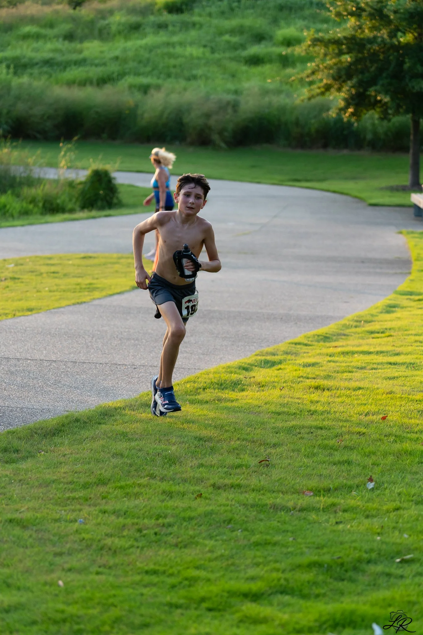 A young boy running outdoors on a paved path, shirtless, holding a water bottle, with a girl in a blue top walking behind him, surrounded by green grass and trees, during late evening or early morning.