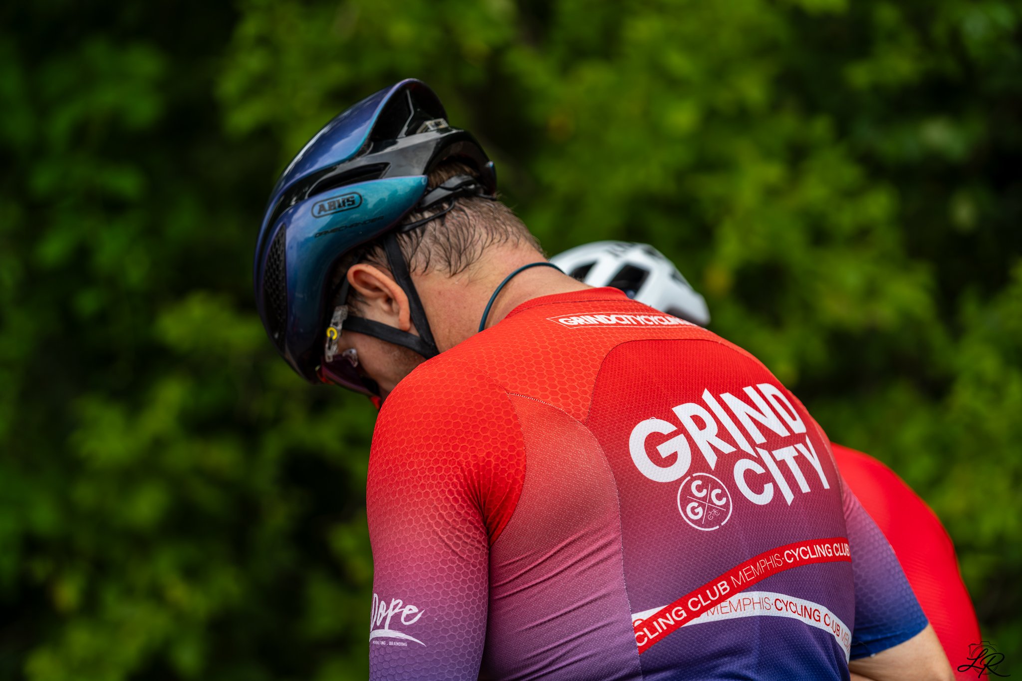 Close-up of a cyclist in a red and purple jersey, wearing a black and blue helmet, with a background of green leaves.