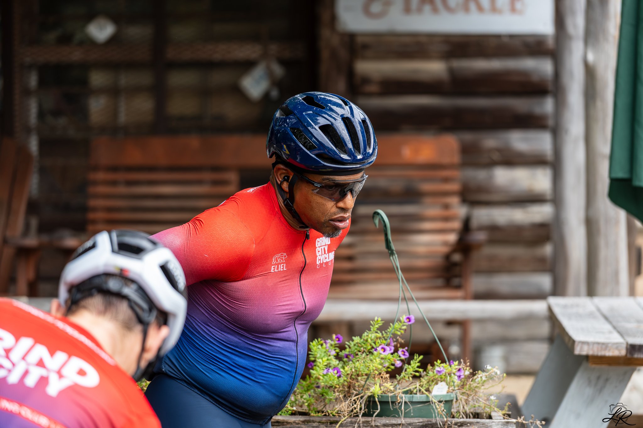 A man wearing a blue bike helmet, sunglasses, and a red cycling jersey is standing near a flower planter, with another cyclist partially visible in the foreground, and a wooden background.