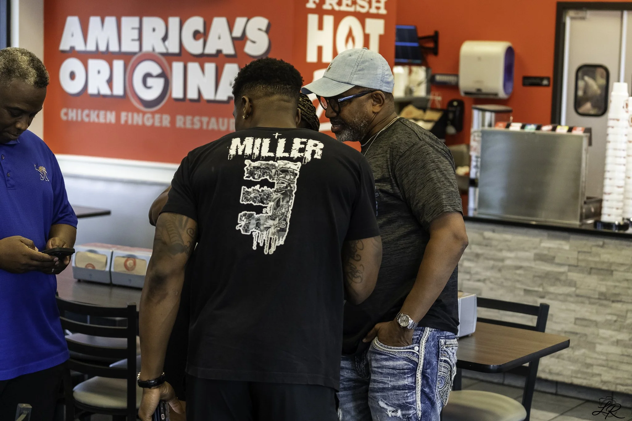 Four men standing together inside a fast food restaurant with a red wall behind them that reads 'America's Original Chicken Finger Restaurant' and 'Fresh Hot'.