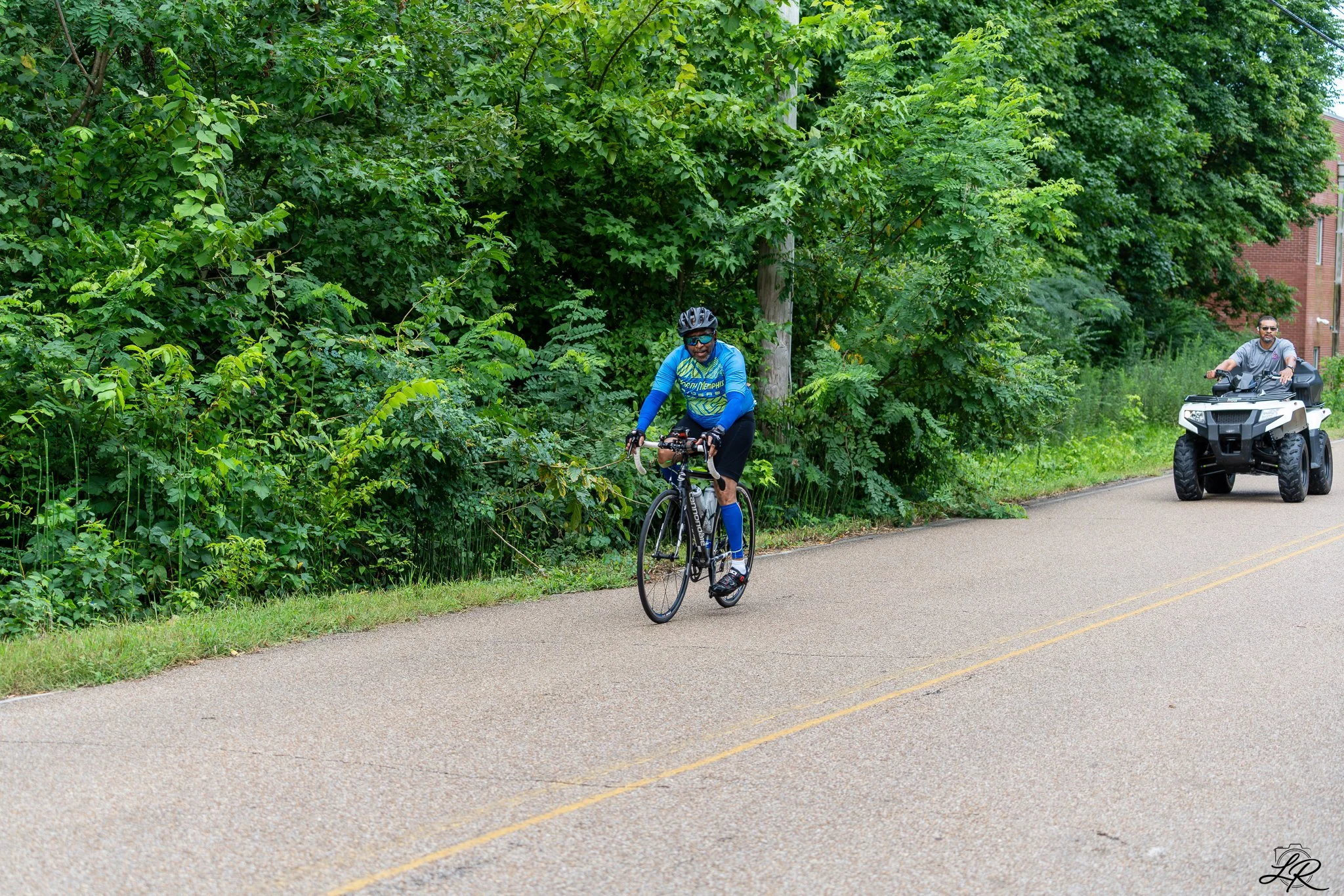 A man riding a bicycle on a paved road with lush green trees on the side, accompanied by a person on a motorized quad bike, in a suburban area with brick buildings in the background.