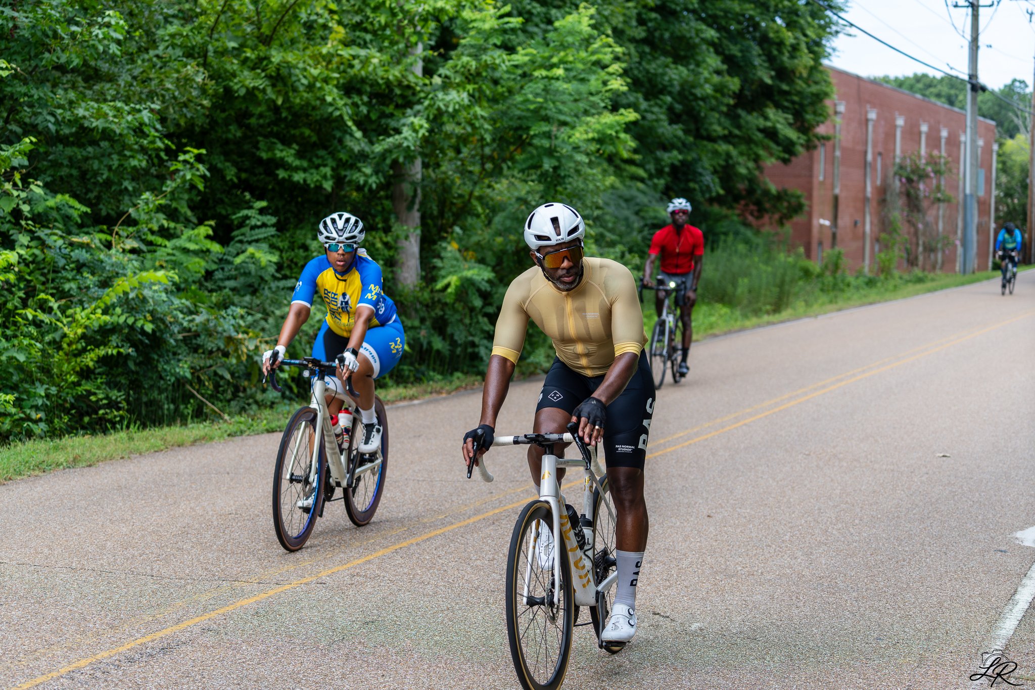 Three cyclists riding on a paved road surrounded by green trees, with a red brick building and utility pole in the background.