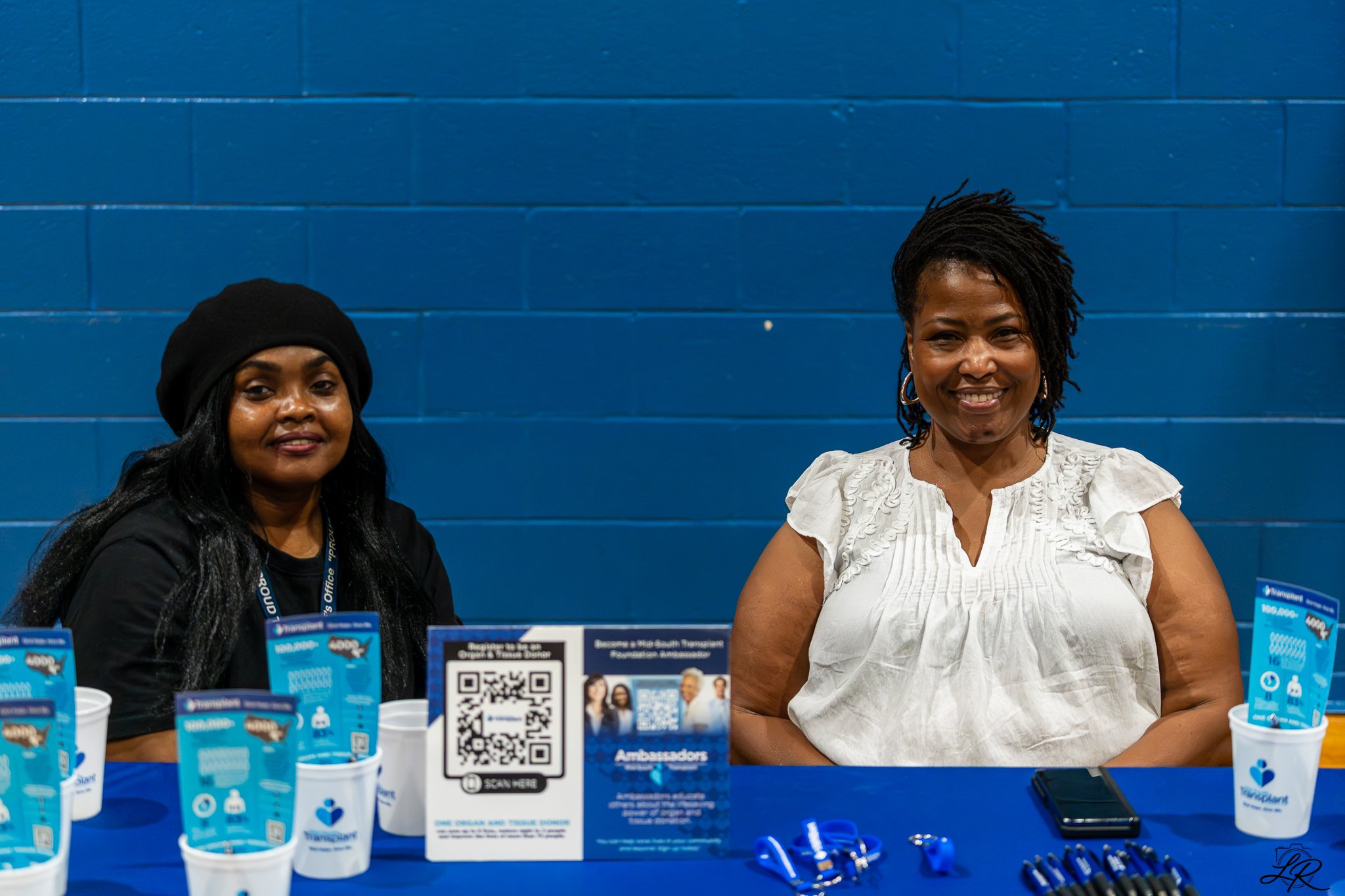 Two women sitting at a table with informational materials and promotional items, smiling, against a blue wall.