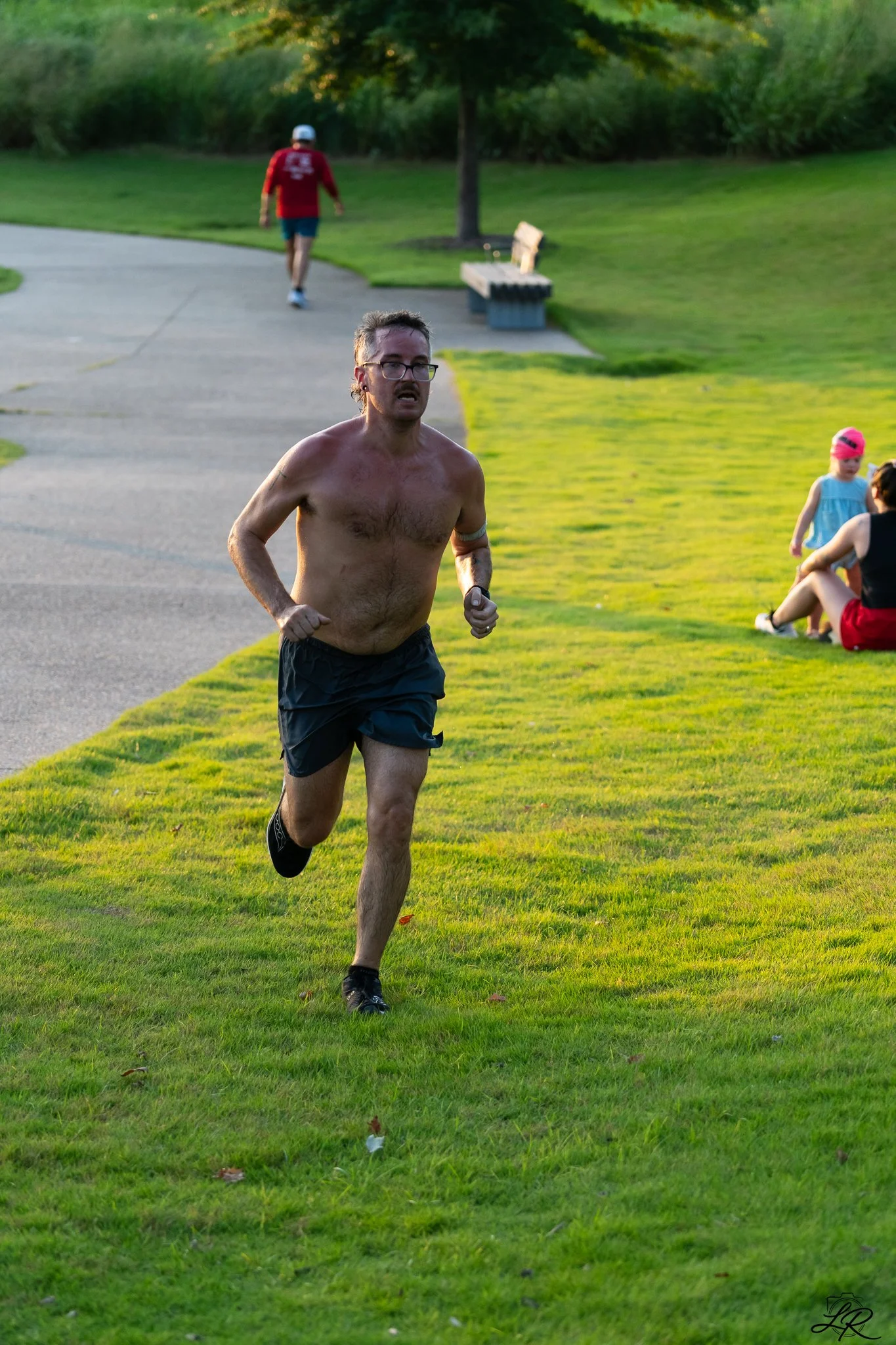A man running shirtless on a grassy park with a tree and a bench in the background, while a person in a pink hat and a woman sit on the grass.