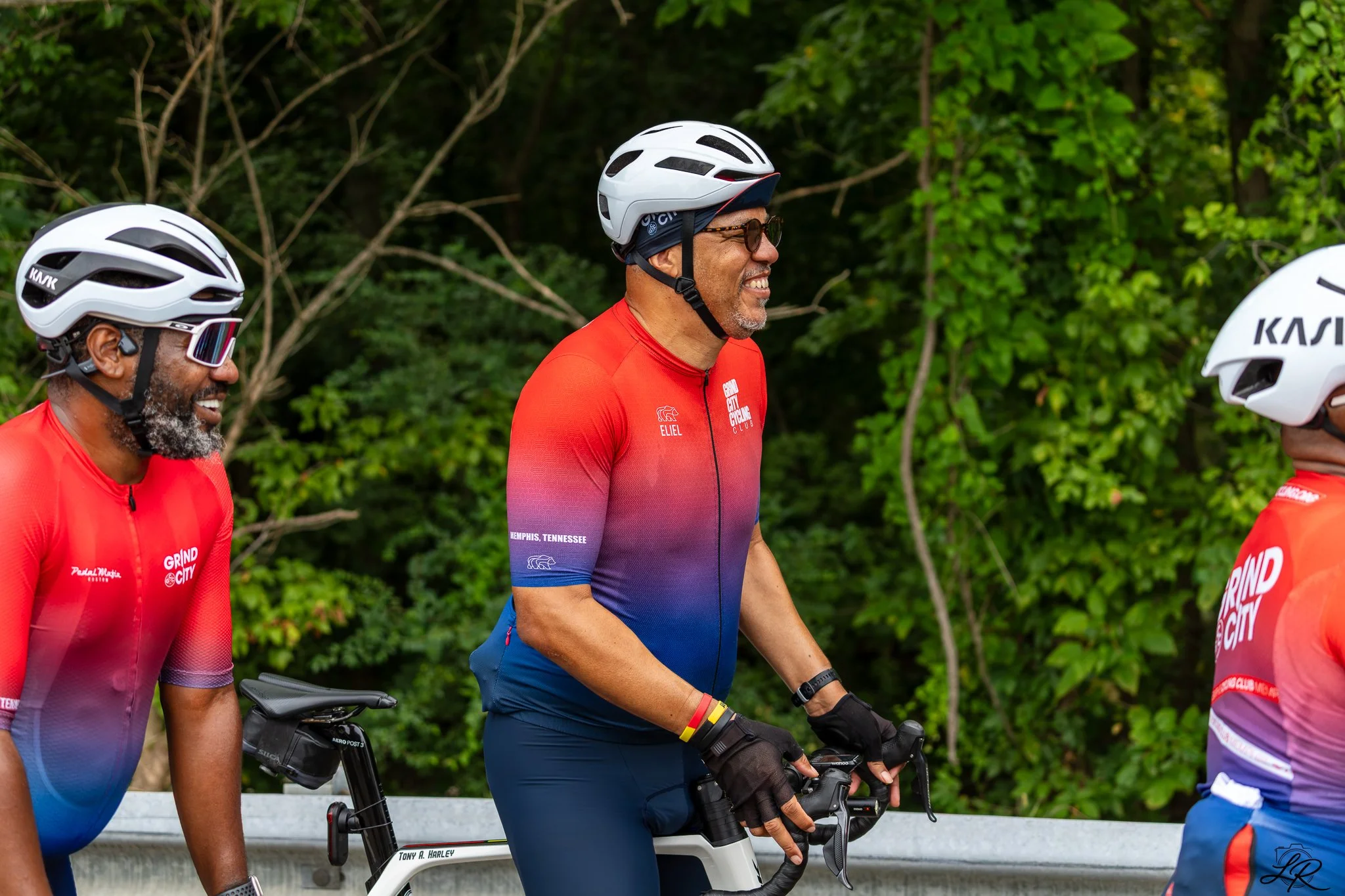 Three people in red cycling jerseys, wearing helmets and sunglasses, riding bicycles on a road with lush green trees in the background.