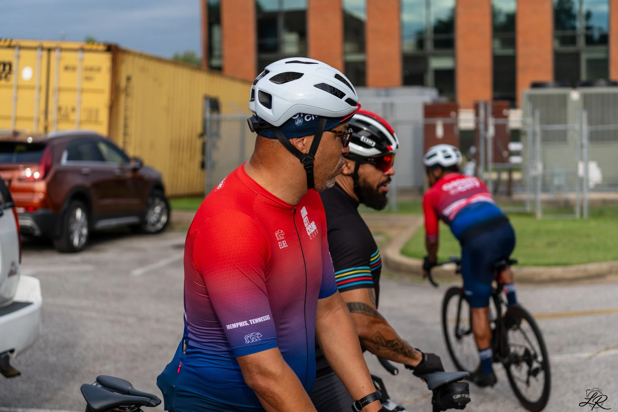 Group of cyclists, including a man wearing a white helmet and red cycling jersey, sitting on their bikes outdoors near a parking lot with cars and a building in the background.